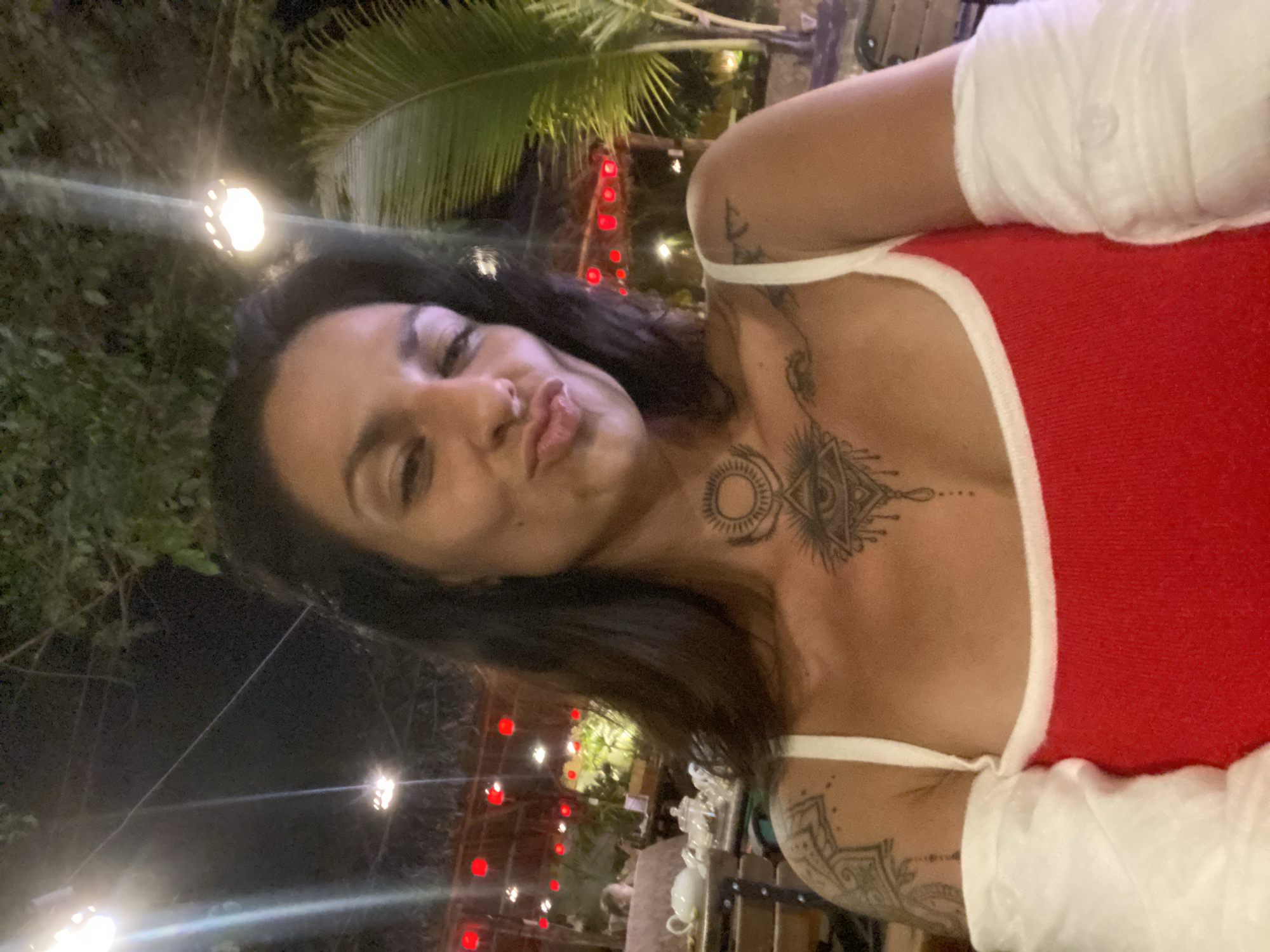 A woman with dark hair and tattoos on her chest and arms is taking a selfie at night. She is wearing a red tank top with white trim and her left arm is bandaged. In the background, there are trees, red lanterns, and palm trees, suggesting an outdoor setting. The presence of teacups and glassware on a table to the left indicates she may be at a restaurant or cafe. The overall ambiance suggests a tropical or subtropical location with decorative lighting. The provided context places the scene in Phu Thuy Ward, Vietnam.
