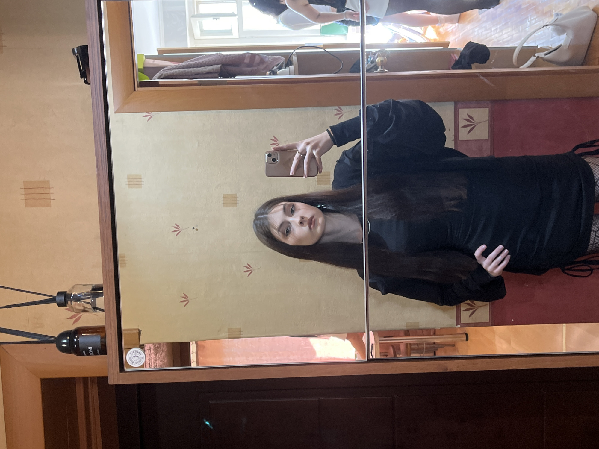 An indoor mirror selfie features a young woman, likely in her late teens or early twenties, taking her own picture. The image is taken in a residential setting, possibly a bedroom or hallway, in Saint Petersburg, Russia.
The woman is reflected in a large, two-panel mirrored wardrobe or cabinet with a light brown wooden frame. She has long, dark brown hair and is wearing a dark, possibly black, long-sleeved top or dress with some volume or detail on the shoulders. She holds a light gold/rose gold smartphone in her right hand, aiming it towards the mirror to capture the selfie. Her expression is neutral as she looks directly at the camera.
The wall beside the mirror is covered in light cream/beige wallpaper featuring subtle patterns of small red-orange leaf clusters and abstract horizontal lines. Two reed diffusers are visible on a ledge next to the mirror; one is clear glass, and the other is amber-colored with the word "AROMI" visible on its label. A circular sticker, partially showing the word "comfort," is affixed to the lower right corner of the left mirror panel. Below the mirrored sections, there is a dark wooden panel.
The reflections in the mirror reveal more of the room: through the right mirror panel, a window with white frames suggests bright daytime natural light. On a shelf inside the wardrobe (reflected in the mirror), folded towels (purple/grey), a white handbag, and a small gold figurine are visible. Another person's lower body, wearing light-colored clothing and dark footwear, is partially reflected in the upper left corner, suggesting they are also in the room. The overall impression is a well-lit, contemporary indoor space.