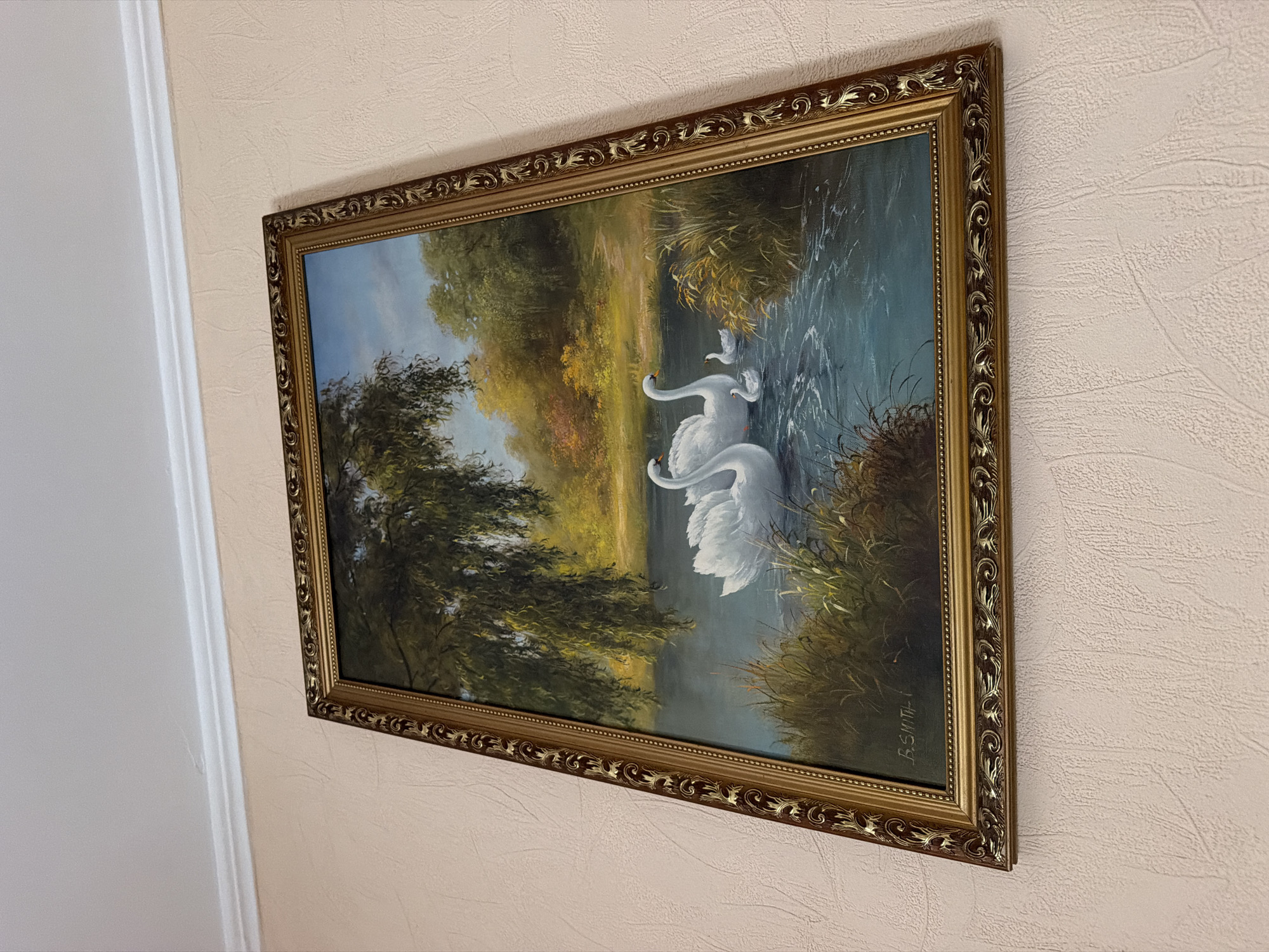 The image displays a painting of a serene natural landscape featuring a family of swans on a body of water. The painting is framed with an ornate, gold-colored frame and hangs on a textured, light-colored wall, below a white ceiling cornice. The painting depicts a lake or river surrounded by lush green trees and foliage, with a path visible in the background. Two adult swans and two cygnets are swimming in the water. The overall impression is one of tranquility and natural beauty. The context provided states that this image is from Mariupol, Ukraine, however, there are no discernible landmarks or contextual elements within the painting or the surrounding environment that would specifically identify its location within the city of Mariupol.