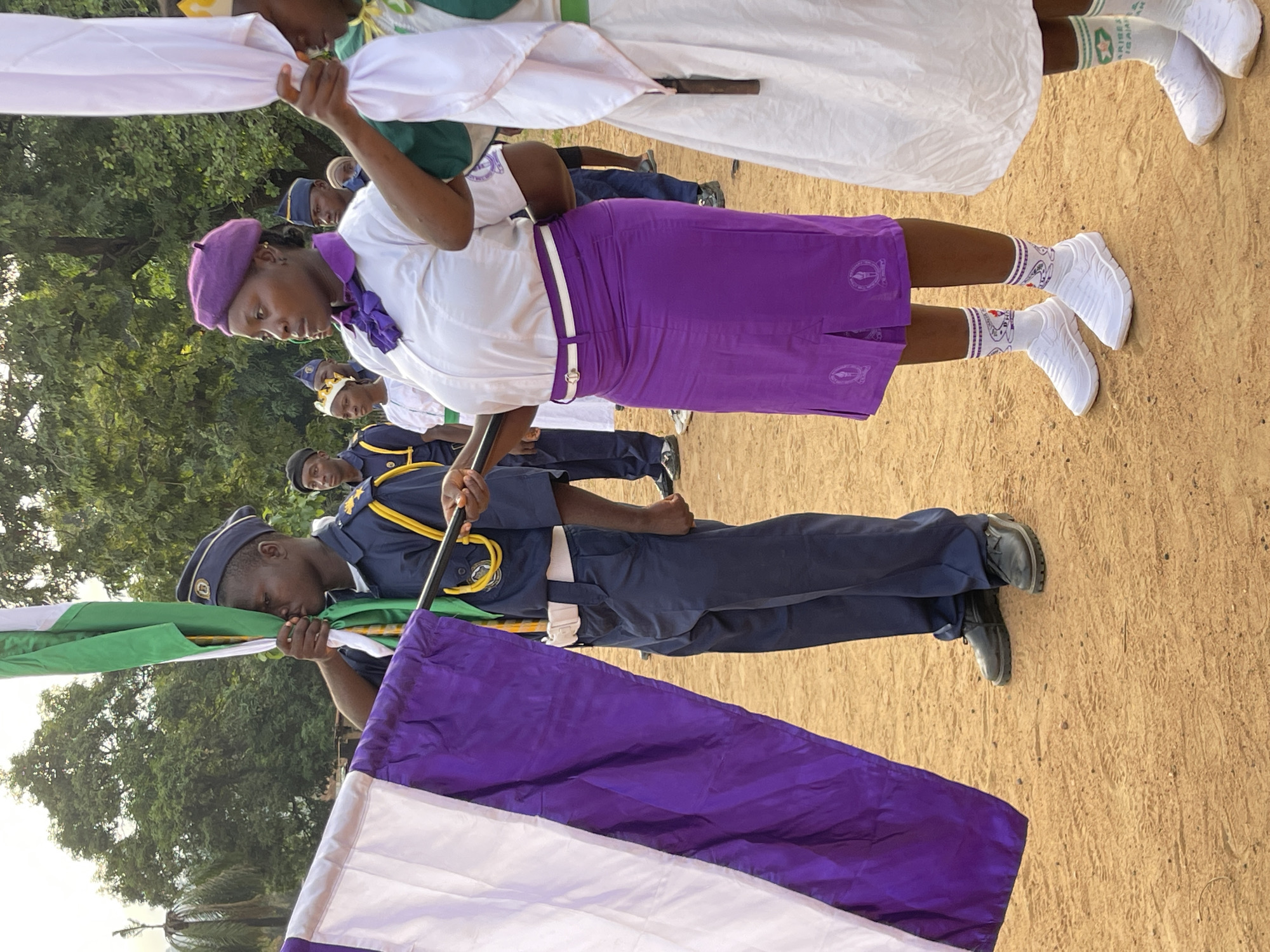 The image shows a group of individuals in Tabu, Nigeria, assembled outdoors. The ground is bare earth. In the foreground, a person in a dark blue uniform, possibly military or a similar service, holds a pole with a flag composed of white, green, and purple stripes. The person appears to be saluting the flag. Other members of a group stand behind them, some in purple skirts and white shirts, and others in white dresses with green accents. All seem to be participating in a formal event or parade. Some of the group are holding white cloths or flags.