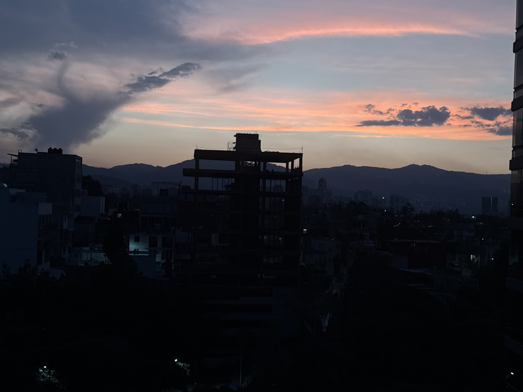 The image depicts a cityscape at dusk in Mexico City, Mexico. In the foreground, the dark silhouettes of buildings and trees are visible. A partially constructed building with a skeletal framework stands prominently in the center of the image. In the background, the silhouettes of mountains are visible against the sky. The sky is a mix of colors, with shades of blue, gray, and pink. The lighting suggests it is either early morning or late evening. There are no people visible in the image.