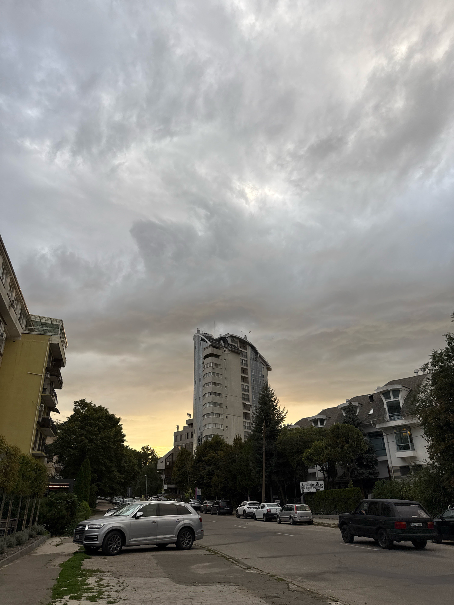 The image depicts a street scene in Chișinău, Moldova, under a cloudy sky with a gradient of yellow to gray. The focus is on a street with several parked cars and a black Range Rover driving away from the camera. In the foreground, a silver Audi Q7 is parked on the left side of the street. To the left of the street, there is a sidewalk with some greenery and a yellow building with balconies. In the background, a tall, modern building with a curved facade stands prominently, flanked by trees. To the right, a white building with a distinctive mansard roof and dormer windows is visible, also surrounded by trees. The overall atmosphere suggests an overcast day in an urban environment.