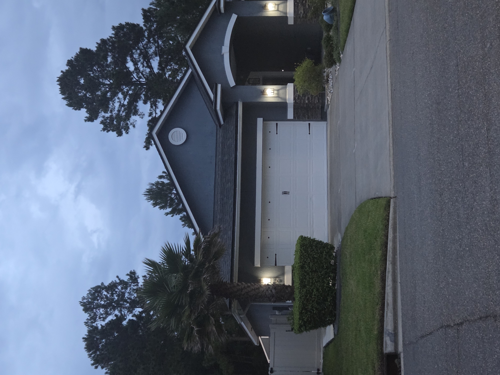 A front exterior view of a single-story, gray stucco house with a white garage door and a covered entryway. The house has a dark gray roof and is surrounded by mature trees and well-maintained landscaping. A palm tree stands to the left of the garage, and a neatly trimmed green hedge is planted in front of it. Two outdoor lights illuminate the garage and the entryway. The driveway is paved with concrete, and the street in front of the house is asphalt. The sky is cloudy and appears to be twilight. The location is the city of Unknown, Unknown.