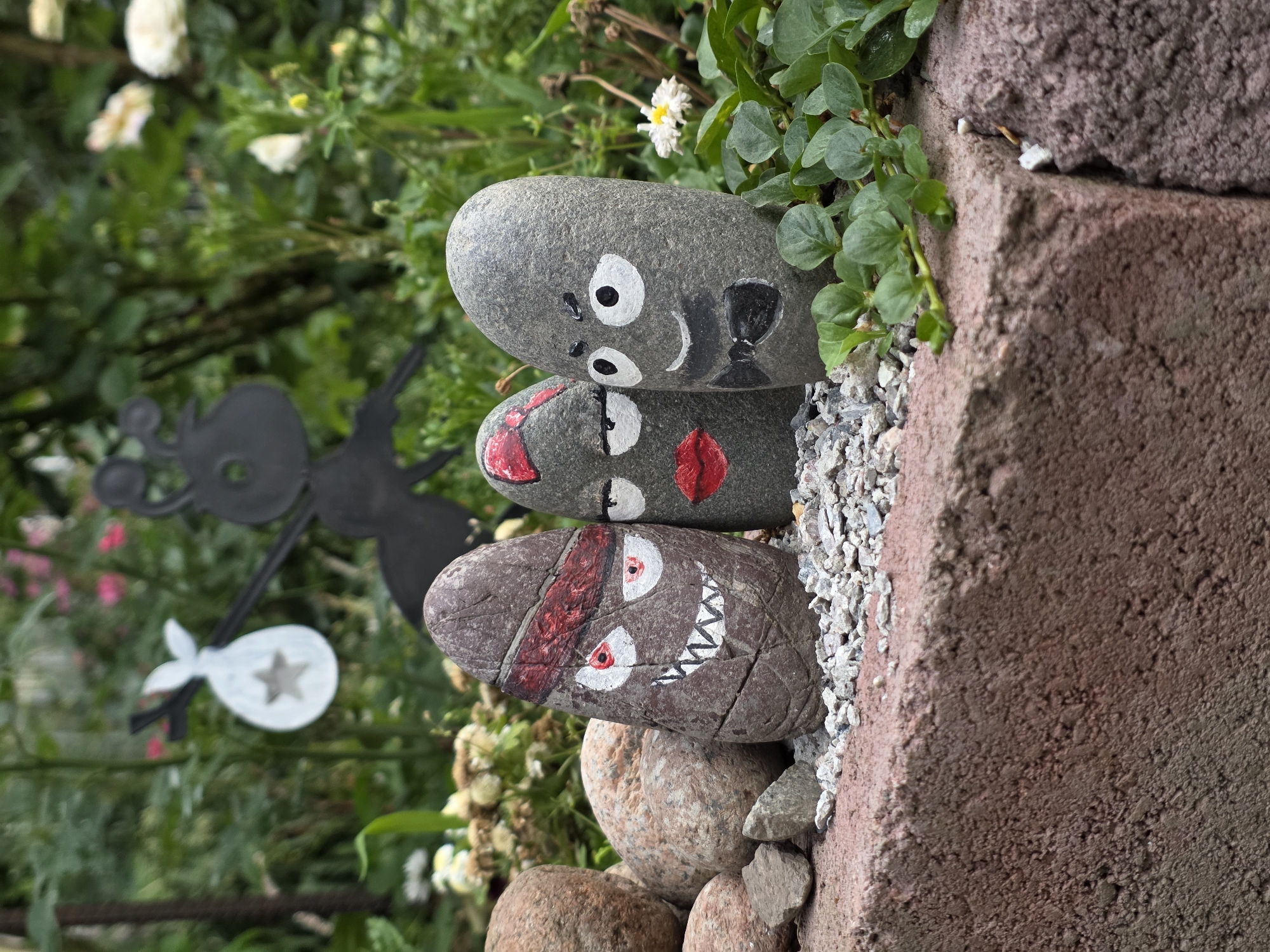 Three painted stones are arranged in a garden bed. The stone on the left is dark brown with a red headband, white eyes with red irises, and sharp white teeth. The middle stone is gray with a red bow-shaped mouth and black eyes. The stone on the right is gray with white eyes, black eyebrows, and a black bowtie. Behind the stones, a black metal silhouette of an ant carrying a bag is visible. The garden bed contains small gray gravel, larger stones, and green foliage. In the background, blurred pink and white flowers are visible. The scene is outdoors, likely in a backyard or garden.