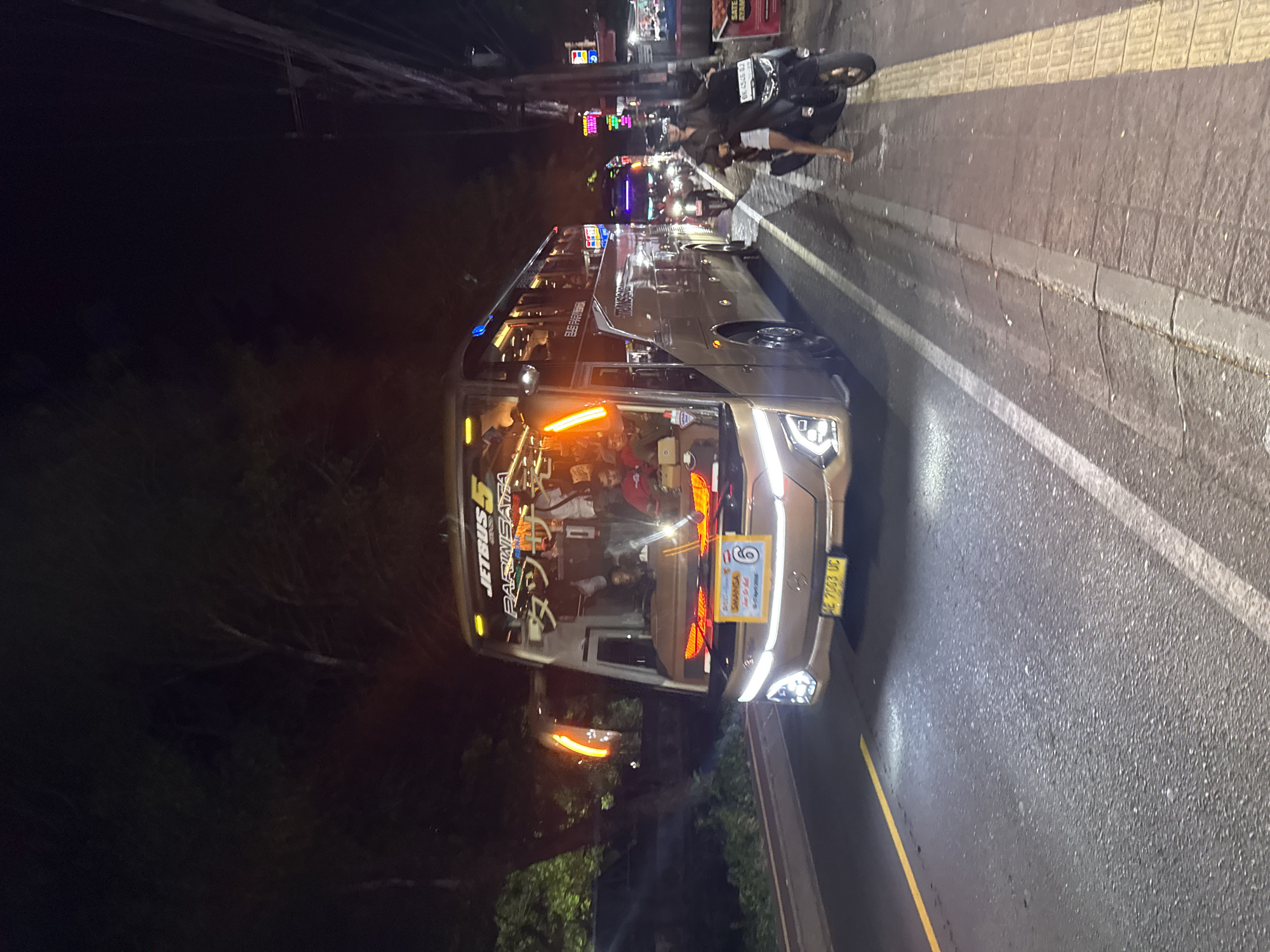 The image depicts a night scene in Ungasan, Indonesia, featuring a large tourist bus parked along the side of a road. The bus, with "JETBUS 5 PARIWISATA" emblazoned on its front, is illuminated by its headlights and internal lights, revealing occupants inside. A motorcycle is parked beside the bus. The background consists of city lights and blurred street activity, suggesting a moderately busy urban environment. The road surface is paved, with a painted yellow line marking its edge.