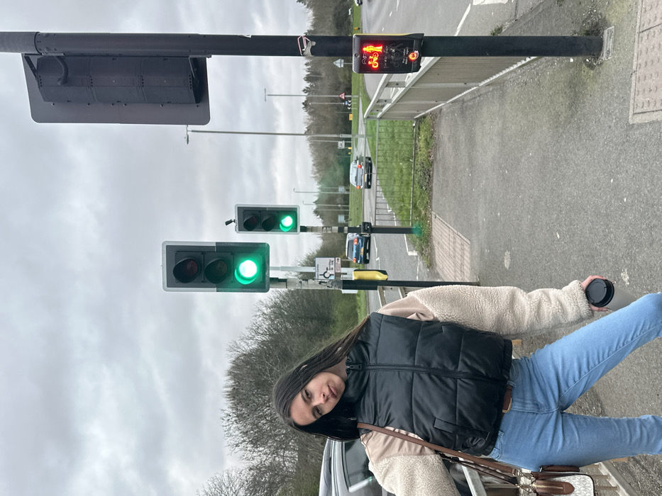 The image depicts an outdoor urban road environment under an overcast sky, specifically at the A6003/A43 Phoenix Parkway Roundabout in Corby, United Kingdom.
A female individual is centrally positioned in the foreground, standing on a paved sidewalk. She has long, dark hair parted in the middle and is looking towards the camera with her head tilted slightly. She wears a dark-colored puffer vest over a beige long-sleeved top with contrasting cream-colored fleece sleeves, light blue jeans, and a brown belt. A brown shoulder bag with a visible "MK" monogram charm hangs over her right shoulder. Her left hand holds a black travel mug.
To the individual's right, a black pole supports a pedestrian/cyclist signal light displaying a red illuminated symbol of a standing person and a bicycle, indicating "do not cross." Adjacent to this pole is a metal guardrail separating the sidewalk from the road. Further into the midground, two vertical traffic light poles are visible, both displaying green lights for vehicular traffic. Below the left traffic light, a small white sign with text and symbols is attached.
The road surface is asphalt, and a grassy median separates traffic lanes. In the background, three vehicles are visible on the road: a dark-colored SUV partially obscured on the left, a blue hatchback, and a silver sedan in the distance, all consistent with left-hand drive vehicles common in the UK. Distant road signs indicating a roundabout (red triangle with circular arrows) and route numbers (A6003, A43) are discernable. The background features bare deciduous trees, suggesting a non-summer season. No direct interactions between individuals are observed; the female individual is posing for the photograph.