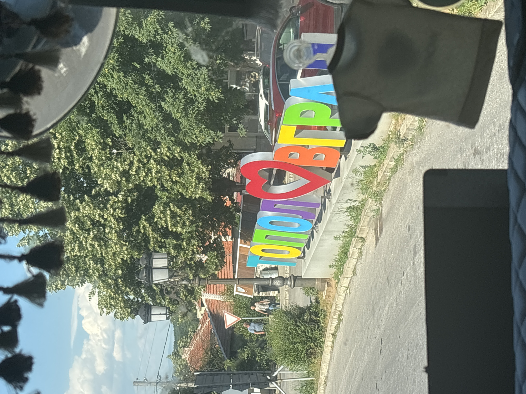 The image shows a street scene in Topolovgrad, Bulgaria, featuring a large, colorful, three-dimensional sign that spells out "ТОПОЛОВГРАД" with a heart symbol in place of the Cyrillic letter "О". The sign's letters are in various bright colors including red, orange, yellow, green, light blue, and purple. In the background, there are trees with lush green foliage, traditional houses with terracotta roofs, and streetlights. Two individuals, a man in a blue t-shirt and a woman in a patterned shirt, are walking on the sidewalk near a yield sign. A red car and other vehicles are parked behind the sign. In the foreground, a dark t-shirt hangs, obscuring a portion of the view. The overall atmosphere suggests a sunny day in a small town.