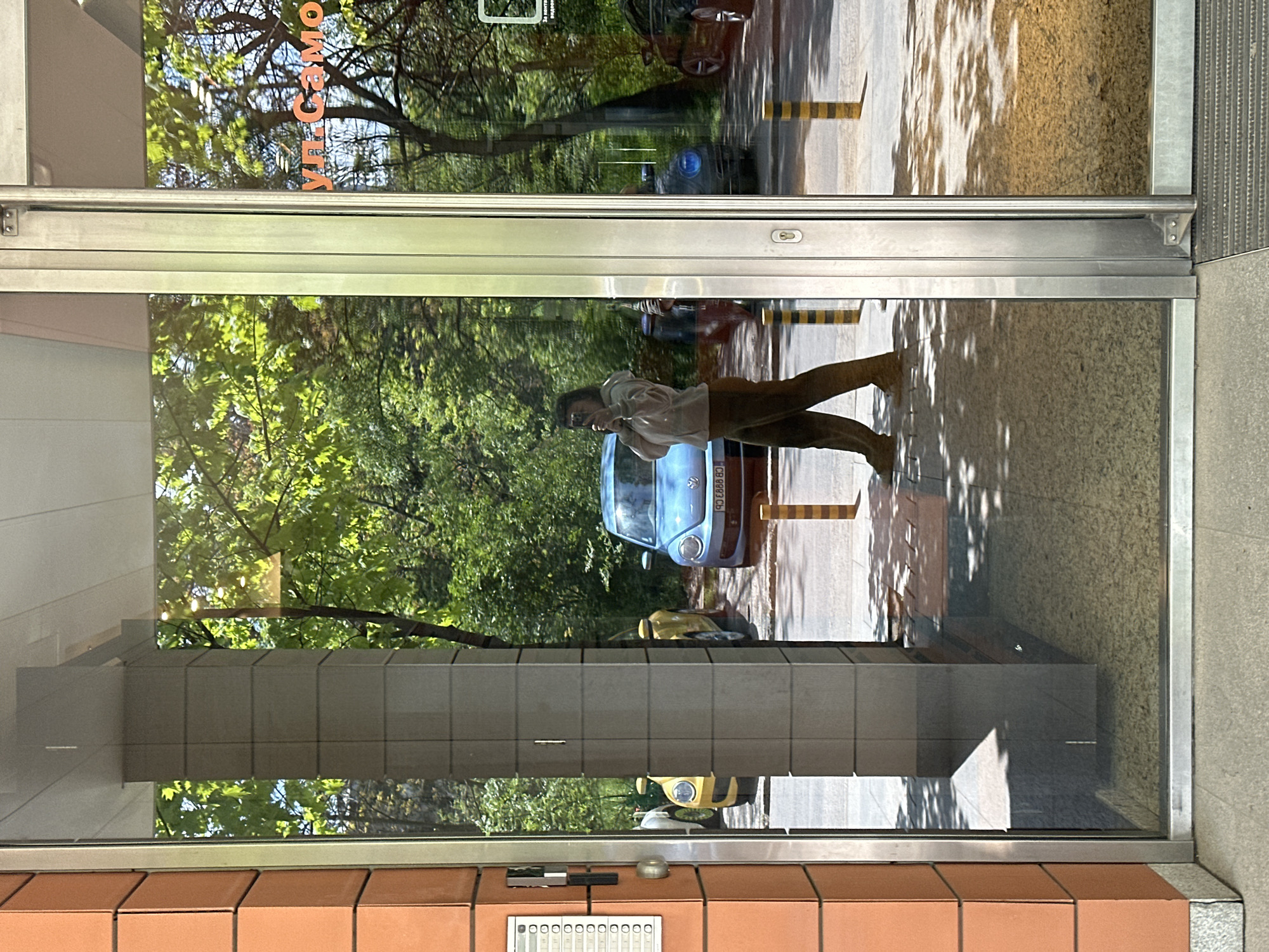 The image is taken from inside a building, looking out through a glass window in Sofia, Bulgaria. The exterior of the building has orange brick tiles. A grey tiled walkway leads to the glass doors. Reflected in the glass is a street with a light blue car, a yellow car and a person. The person is wearing a white shirt, brown pants, and has dark hair, and appears to be walking on the sidewalk. The reflection also shows trees, suggesting an outdoor, urban environment. The reflection also includes the text "VII.CAMO.HK"