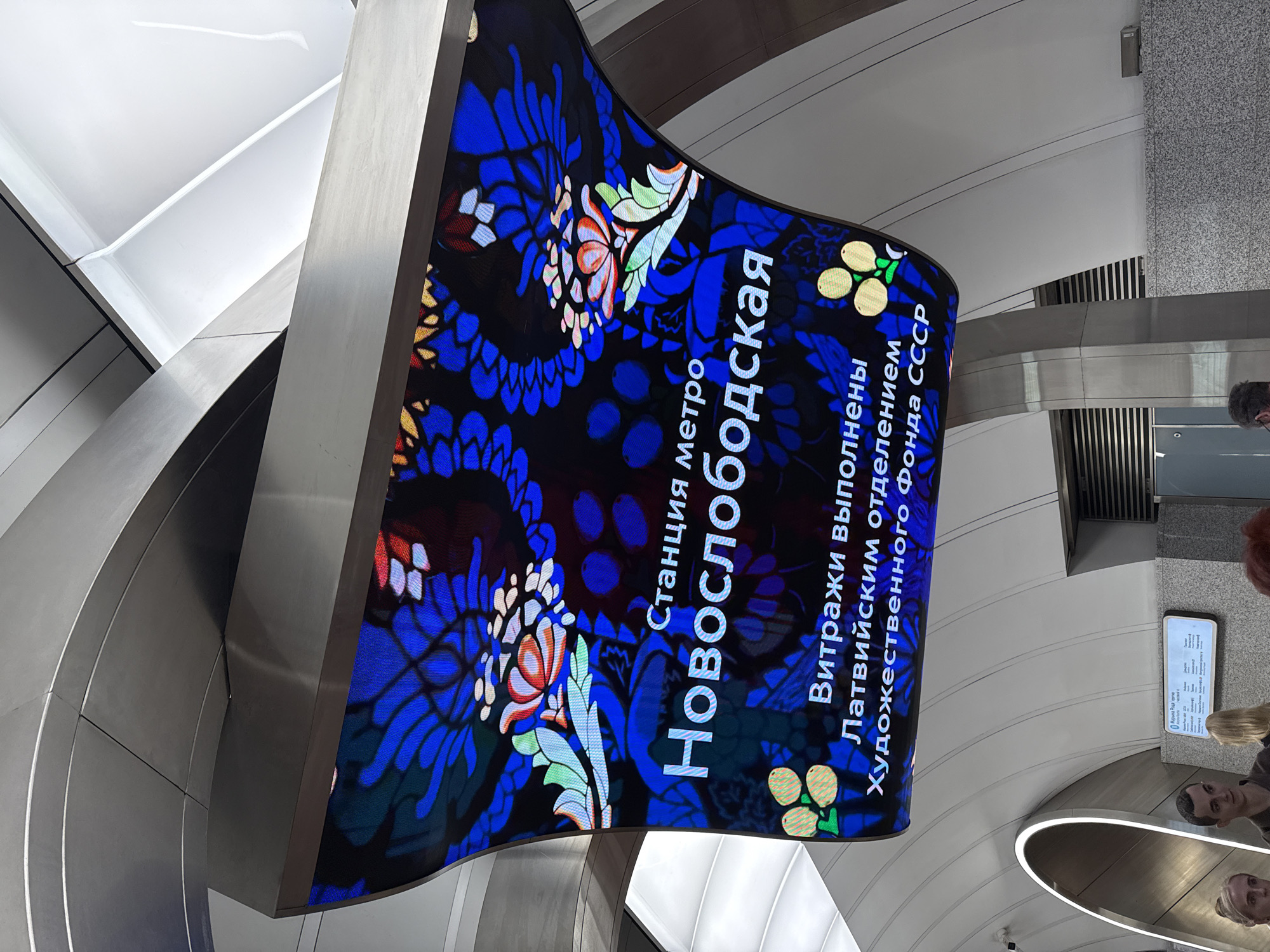 Here is a description of the image:
The image shows a sign in the Novosslobodskaya metro station in Moscow, Russia. The sign is a curved LED display with a floral pattern in shades of blue, black, white, and red. White text on the sign reads "Станция метро Новослободская" (Novosslobodskaya Metro Station) and "Витражи выполнены Латвийским отделением Художественного фонда СССР" (Stained glass windows made by the Latvian Department of the Art Fund of the USSR). The sign is attached to a gray metal structural support. The background shows the curved, white ceiling of the station. Parts of people are visible.