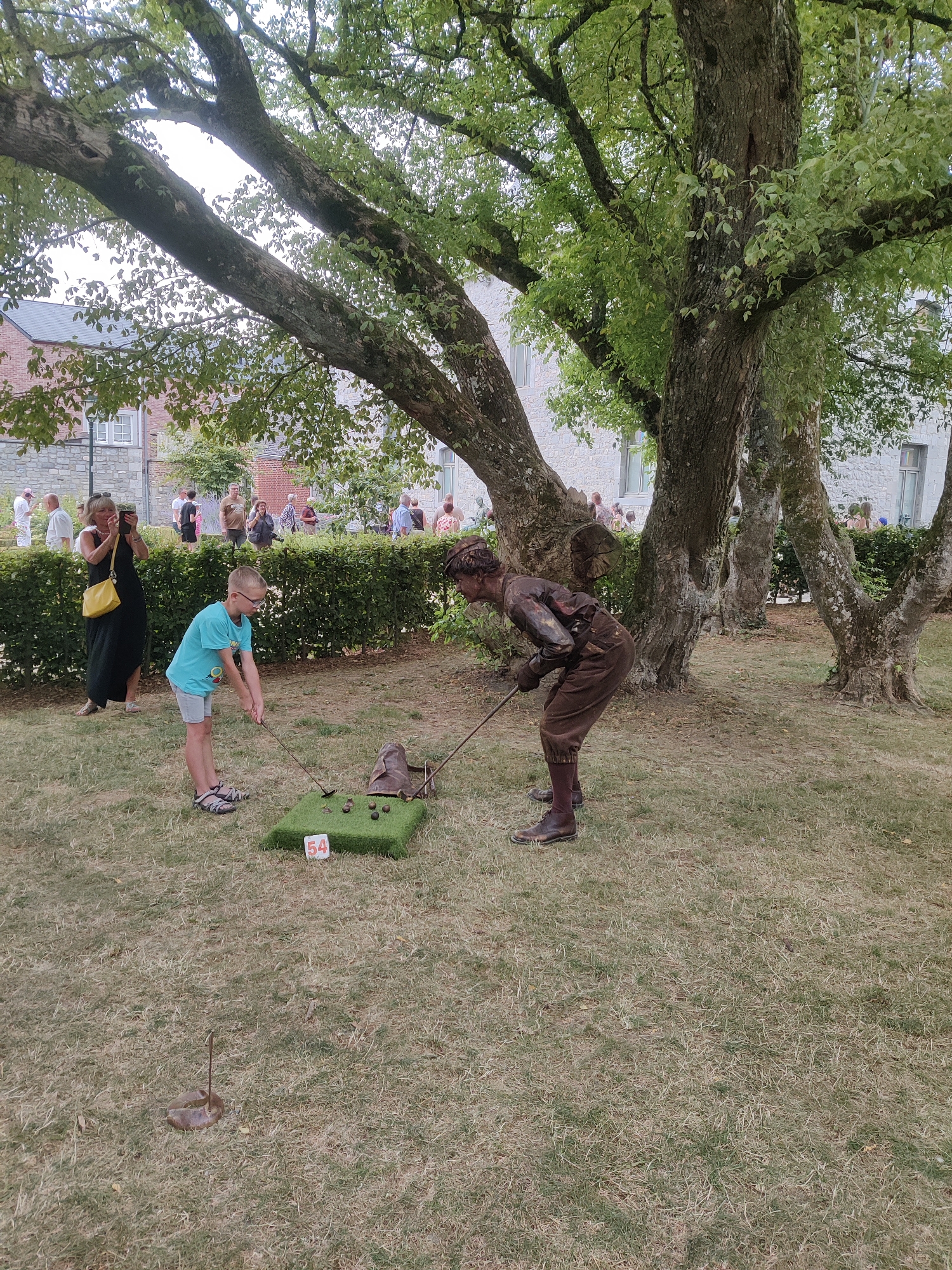 In Marche-en-Famenne, Belgium, two individuals are engaged in a game on a grassy outdoor area. A young boy, wearing a turquoise t-shirt and grey shorts, stands with a golf club, poised to strike a golf ball on a miniature putting green that is marked with the number "54". To his right, a person dressed in brown, body-painted to appear as a statue, is also holding a golf club and appears to be mid-swing. Behind them, a dense green hedge separates the playing area from a gathering of people observing the event. In the background, buildings with stone facades and windows suggest an urban setting, partially obscured by the lush green canopy of mature trees. The ground is covered in dry grass, with a metal stake and horseshoe visible in the foreground. The scene captures a moment of playful activity within a public space.