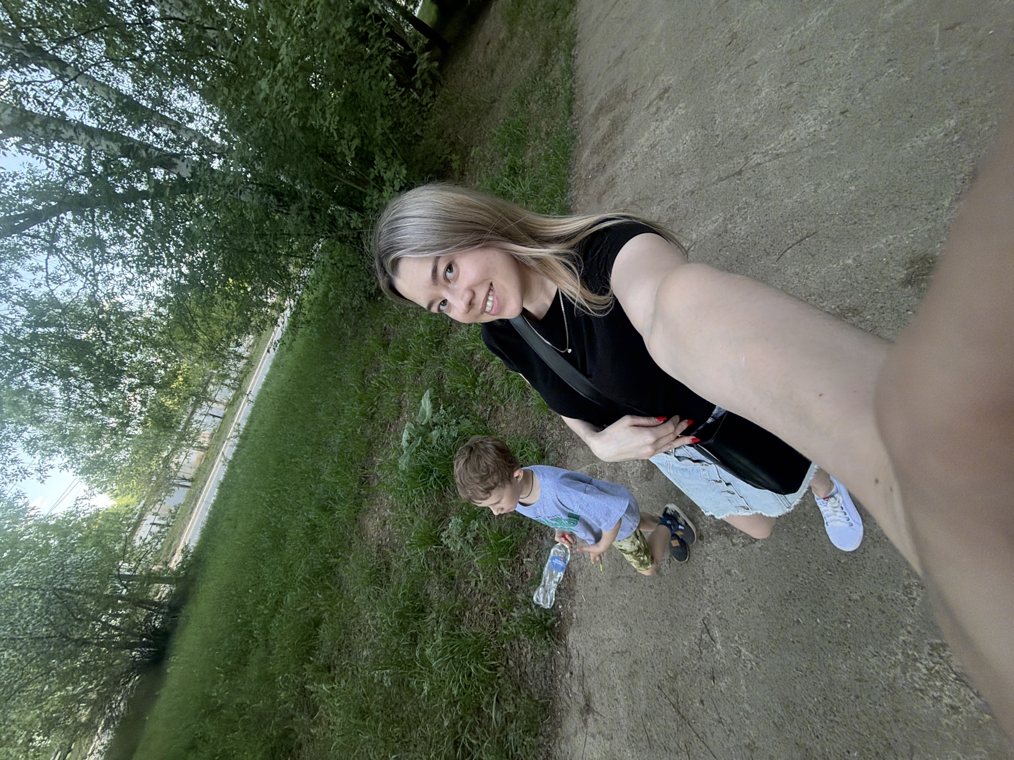 A woman is taking a selfie with a young boy on a dirt path. The woman has blonde hair, wears a black shirt, and has a black purse strapped across her body. She wears a light blue denim skirt and white shoes. The boy is shorter than the woman and wears a light blue shirt, camouflage shorts, and sandals. He is holding a water bottle. The path is surrounded by green grass and trees. The scene appears to be outdoors, potentially in a park. The location is Chaikovsky, Russia.