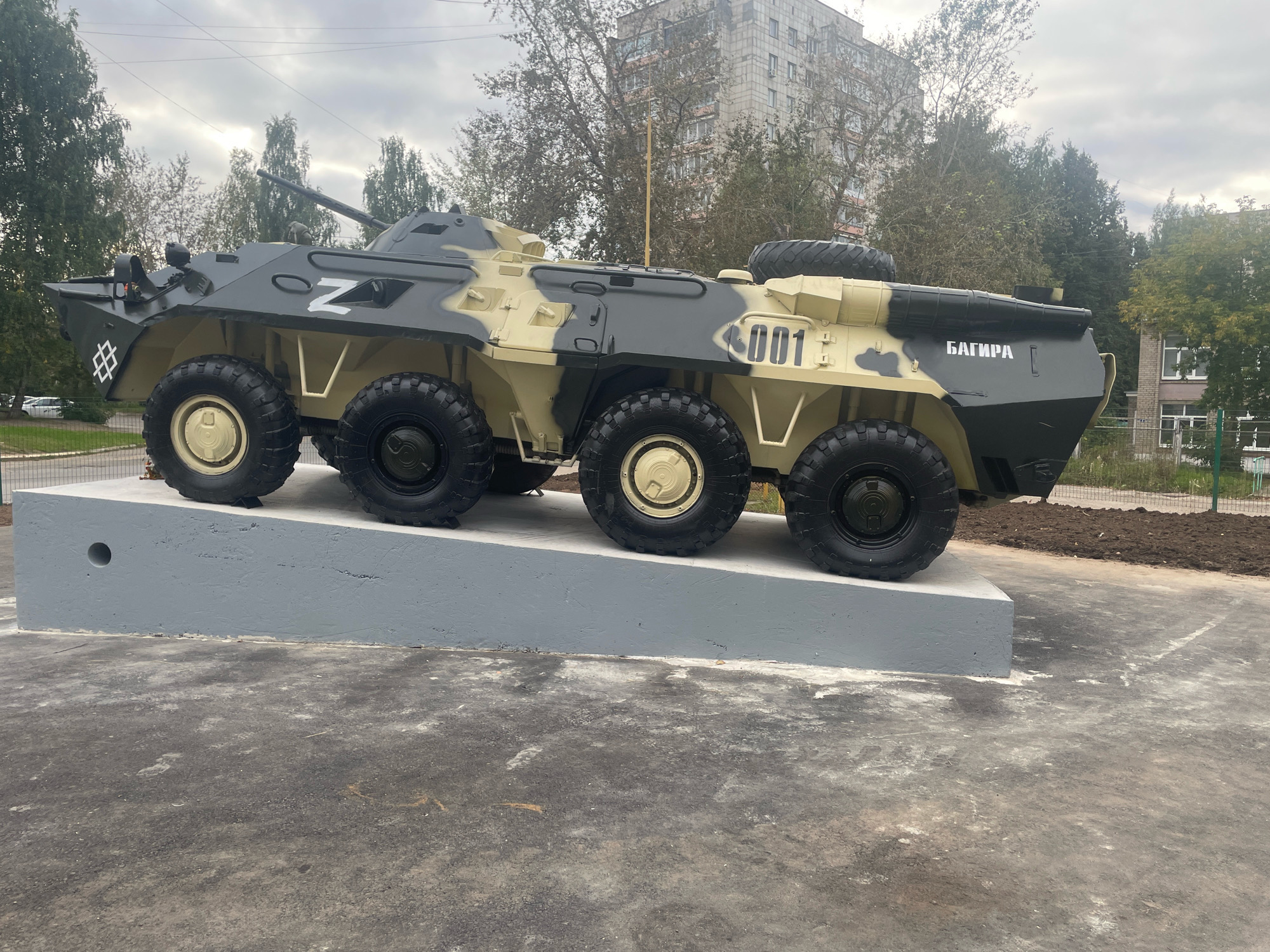 An outdoor, low-angle, full shot captures a six-wheeled armored personnel carrier (APC) painted in a camouflage pattern of dark green, tan, and black. The APC is elevated on a light gray concrete platform that slopes down to the right. The APC has a turret on its roof and a spare tire mounted on its rear. Markings on the vehicle include a white "Z" symbol on the left side, the number "001" in white on the right side, and the Cyrillic word "БАГ ИРА" (BAGIRA) also in white on the right rear. In the background, a multi-story residential building with windows is visible, along with trees that have green and yellow leaves. The sky is overcast and cloudy. The ground in front of the platform is paved with asphalt. The scene is located in the city of Chaikovsky, Russia. No people are present.