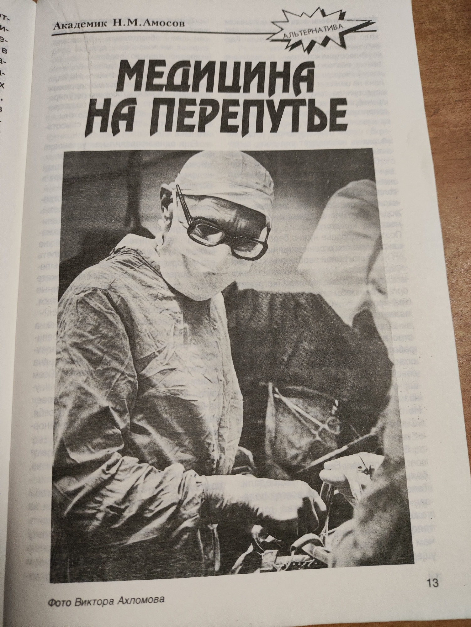The image is a black and white photograph of a newspaper page. At the top, centered, is the text "Академик Н.М.Амосов" (Academician N.M. Amosov). To the right of this, in a jagged, starburst-like graphic, is the word "АЛЬТЕРНАТИВА" (Alternative). Below this, in large, bold, capital letters, is the title "МЕДИЦИНА НА ПЕРЕПУТЬЕ" (Medicine at a Crossroads).
The main subject of the photograph is a surgeon in scrubs and a surgical cap, wearing a surgical mask and glasses, performing surgery. The surgeon is in the foreground, looking down at the surgical field. Surgical instruments are visible on a tray in front of the surgeon. Another person in surgical attire is partially visible in the background.
At the bottom left of the page, there is text that reads "Фото Виктора Ахломова" (Photo by Viktor Akhlomov). At the bottom right, the page number "13" is visible.
There is no information in the image that indicates a location in the city of Izhevsk, Russia.
