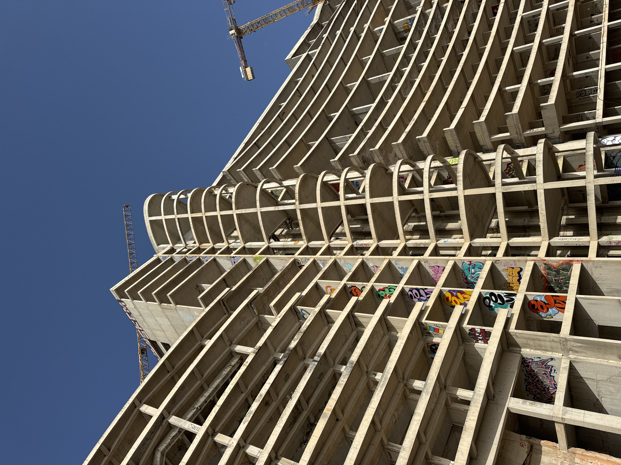 The image shows a low-angle shot of an unfinished concrete building with graffiti on some of the inner walls of the frame. The building is located in Perazića Do, Montenegro. The frame is incomplete, showing the support structures and the lack of exterior walls. A construction crane is visible in the background against a clear blue sky. The building's architecture includes both linear and curved elements.
