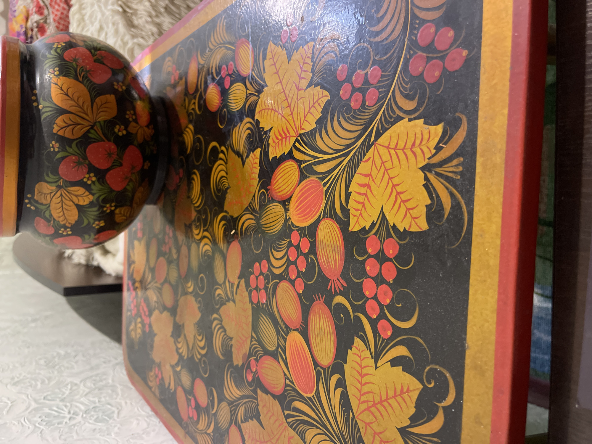 A close-up shows a side view of a black wooden object decorated with colorful Khokhloma painting, featuring yellow and orange leaves, red berries, and spiraling patterns on a glossy black background. The object has a rectangular base with red and yellow trim and a bulbous top also adorned with Khokhloma patterns. The image appears to have been taken indoors, with a white textured surface in the foreground.