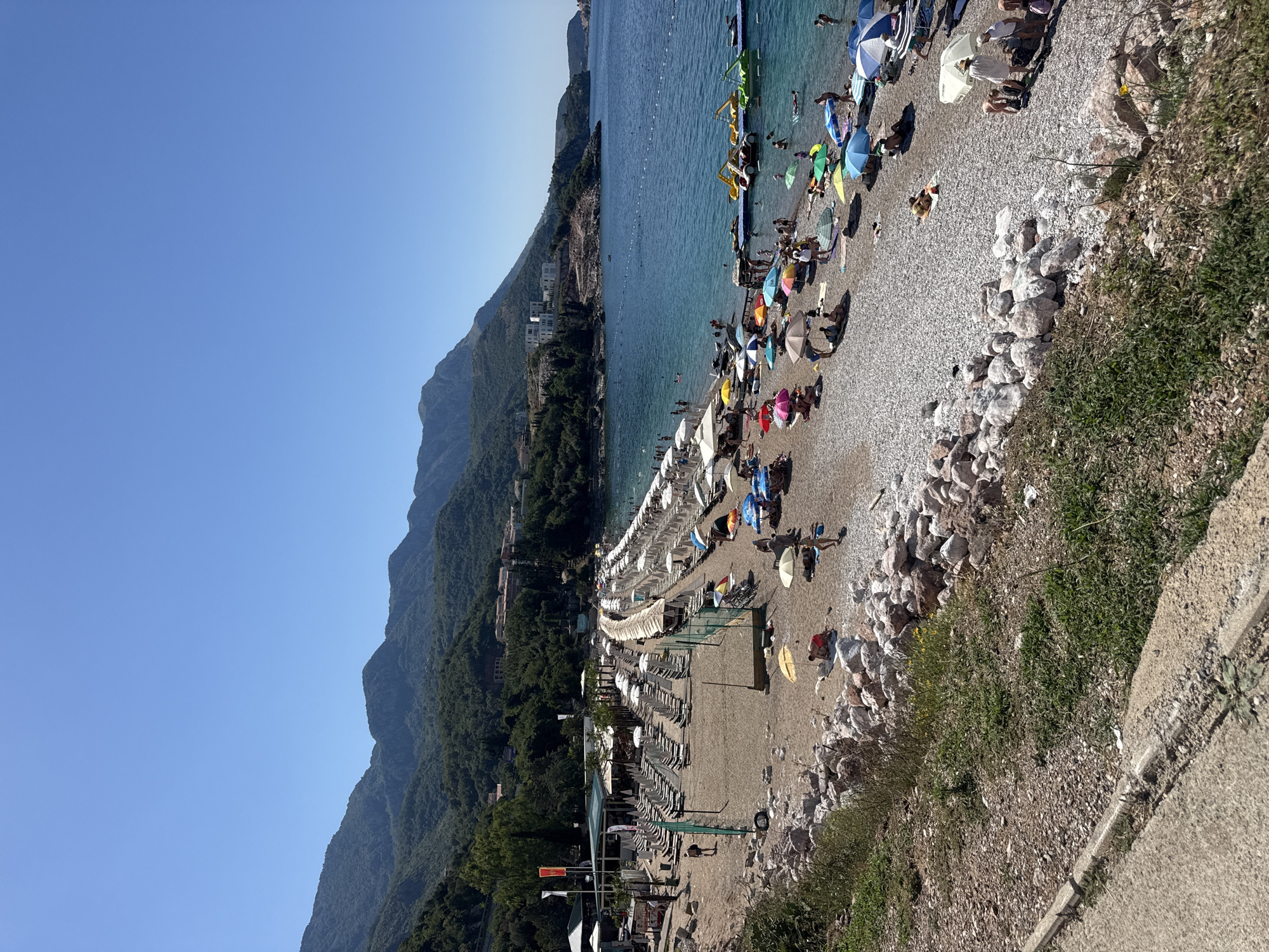 This image depicts a vibrant beach scene in Kamenovo, Montenegro, under a clear blue sky. The expansive beach is covered with pebbles and sand, lined with numerous rows of white lounge chairs and umbrellas. Several beachgoers are scattered across the beach, some sunbathing, others walking, and some gathered under umbrellas. The azure water of the Adriatic Sea laps gently at the shore, with a few individuals swimming or wading. In the water, there are several colorful pedal boats, indicating recreational activities.
The background features a lush, green mountainside that slopes down towards the coastline, dotted with buildings and vegetation. The topography suggests a coastal town nestled against a mountainous backdrop. A paved path or road runs along the foreground, with some greenery and rocks bordering it. The overall impression is a lively and picturesque beach resort on a sunny day.