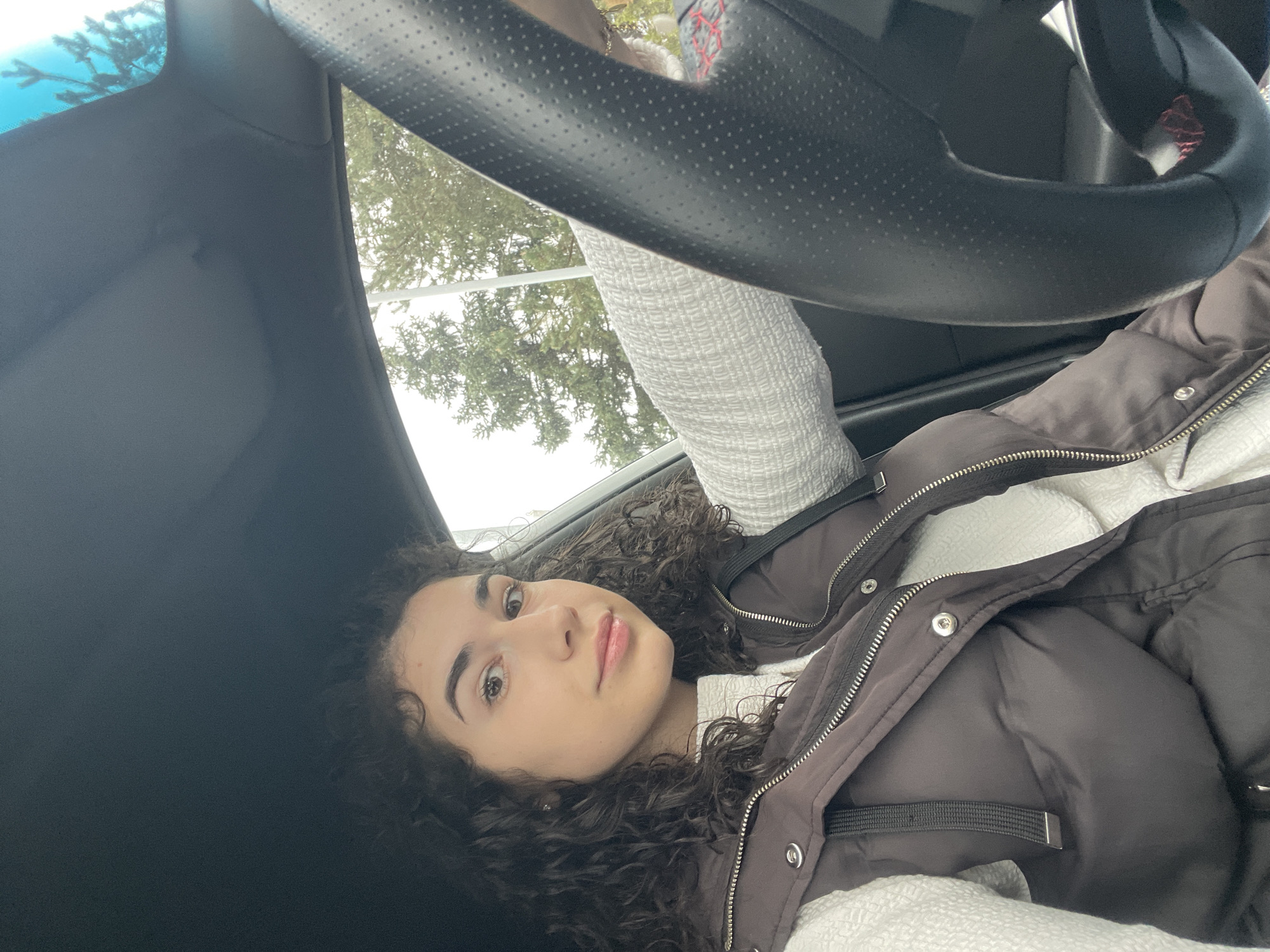 The image shows a young woman positioned inside a car, looking directly at the camera. The image is rotated approximately 90 degrees counter-clockwise. She has dark, curly hair, fair skin, and a calm, neutral expression with subtle pink lip color.
She is wearing a dark brown or grey puffer jacket featuring silver zippers and snaps, layered over a white, textured long-sleeved top. The setting appears to be the interior of a vehicle, with a black, perforated steering wheel prominently visible in the upper right quadrant of the rotated image. Parts of the car's dark fabric headliner are visible in the upper left. A white textured material, possibly a blanket or part of her clothing, is also seen near the steering wheel.
Through the car windows, a bright, overcast sky and green trees with dense foliage are visible, suggesting daytime. There are no specific visible cues within the image to confirm the location, but the provided context states it is Sofia, Bulgaria. The primary activity is the woman posing for a photograph. No visible text is present in the media file.