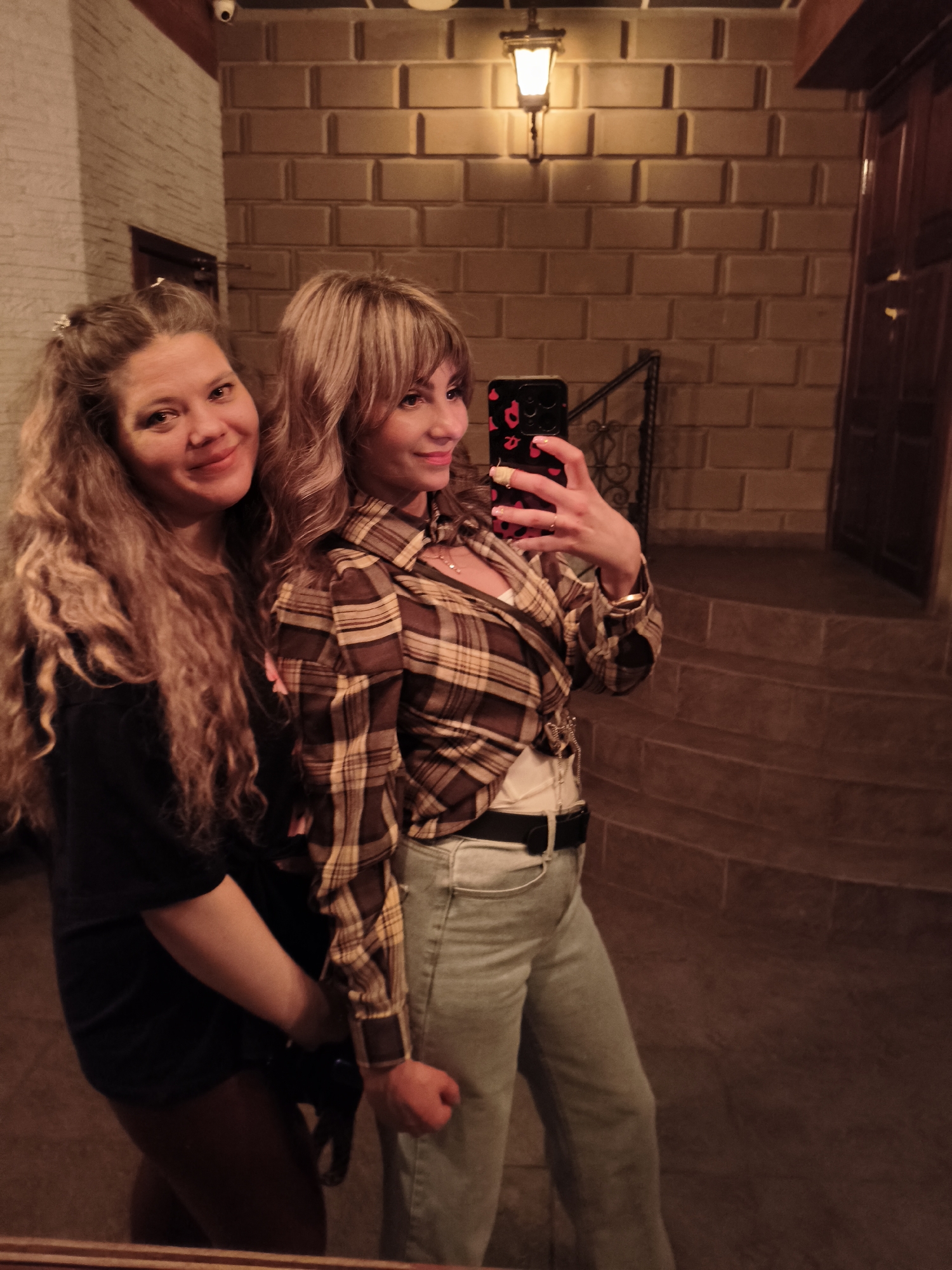 The image features two women posing for a selfie in what appears to be an indoor or covered setting, likely during the evening or night, given the artificial lighting. The background consists of walls made of light brown or beige rectangular bricks or tiles.
On the left, a woman with long, wavy, light brown hair and wearing a dark top smiles warmly towards the camera. Next to her, on the right, a second woman is actively taking the photo with a smartphone, looking at the screen with a cheerful smile. She has lighter, slightly wavy hair with bangs and is dressed in a brown and beige plaid shirt over a white undershirt, light blue jeans, and a black belt. Her smartphone is housed in a black case embellished with red leopard print or abstract red spots.
Notable details in the setting include a traditional wall sconce providing light on the right wall, and a segment of a black wrought iron railing visible behind the selfie-taker. Further to the right, a set of dark stairs ascends towards a closed wooden door. A small security camera is mounted on the ceiling in the upper left corner, suggesting a public or semi-public space. Both women display happy and friendly expressions. No legible text is visible in the image.