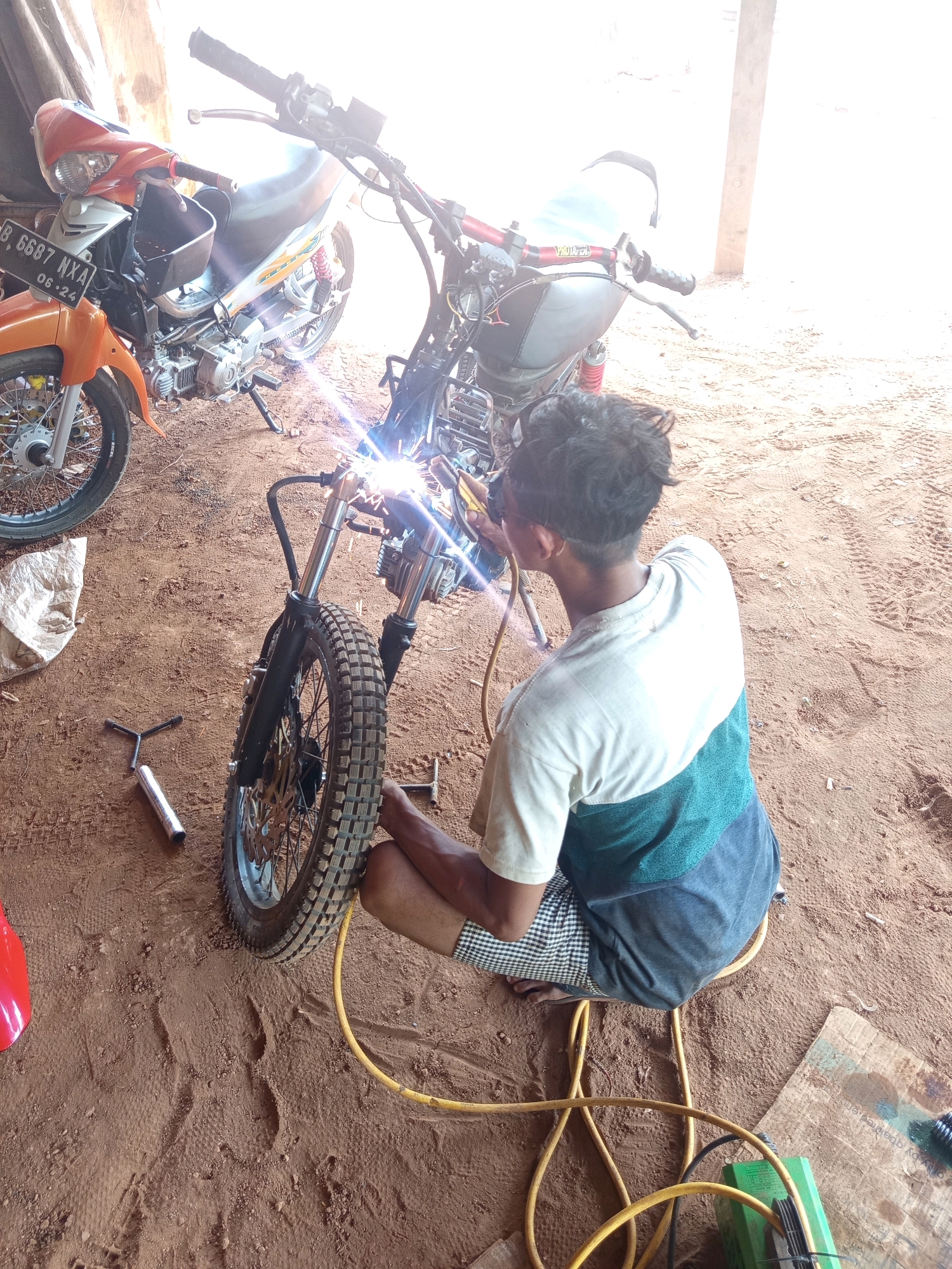 In Karang Sari, Indonesia, a young man is welding a motorcycle frame in an open-air workshop. He is seated on a stool, wearing a t-shirt and shorts, and holding a welding torch. Sparks fly from the point of contact, illuminating the front end of the motorcycle he is working on. A second, partially visible motorcycle, an orange scooter, is parked to the left of the frame. The ground is covered in sandy soil, with tire tracks and scattered debris. In the background, sunlight streams in from an opening, creating a bright, hazy atmosphere.