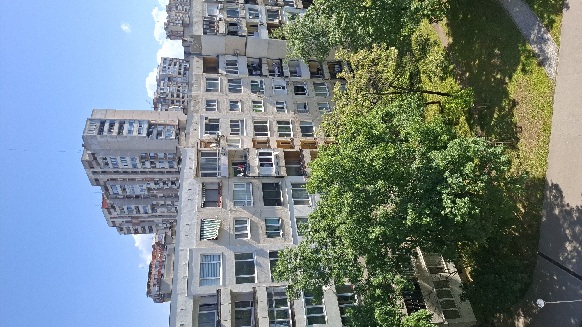 The image shows a residential area in Sofia, Bulgaria, characterized by its dense, Soviet-era architecture. The dominant feature is the block of concrete apartment buildings, with a facade of numerous windows and balconies, indicative of a high-density urban environment. The buildings have a somewhat dilapidated appearance, suggesting the age and the wear of the structure.
The presence of trees with lush green foliage offers a contrast to the concrete, indicating an attempt to incorporate greenery into the urban space.
The sky is blue with few clouds, suggesting clear weather.