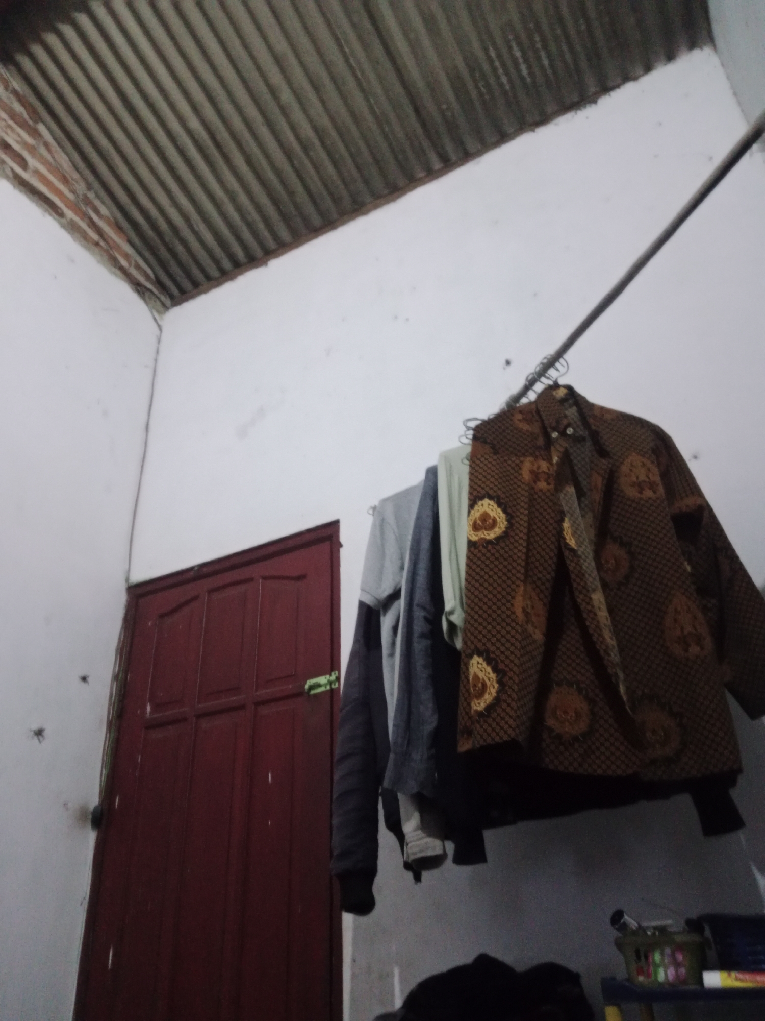 The image depicts an interior scene, likely a room in a residential building in Kejapanan, Indonesia. A dark red door is visible on the left side of the frame, with a light green latch. Above and to the right of the door, clothing items are hanging on a clothesline or rack suspended from a metal rod. The foremost item is a brown patterned long-sleeved shirt or jacket. Behind it, a gray garment and a pale green garment are visible. The ceiling appears to be corrugated metal with visible brickwork at the edge. The walls are painted white and show some signs of wear or staining. The overall impression is of a utilitarian space, possibly a laundry area or a bedroom where clothes are being dried.