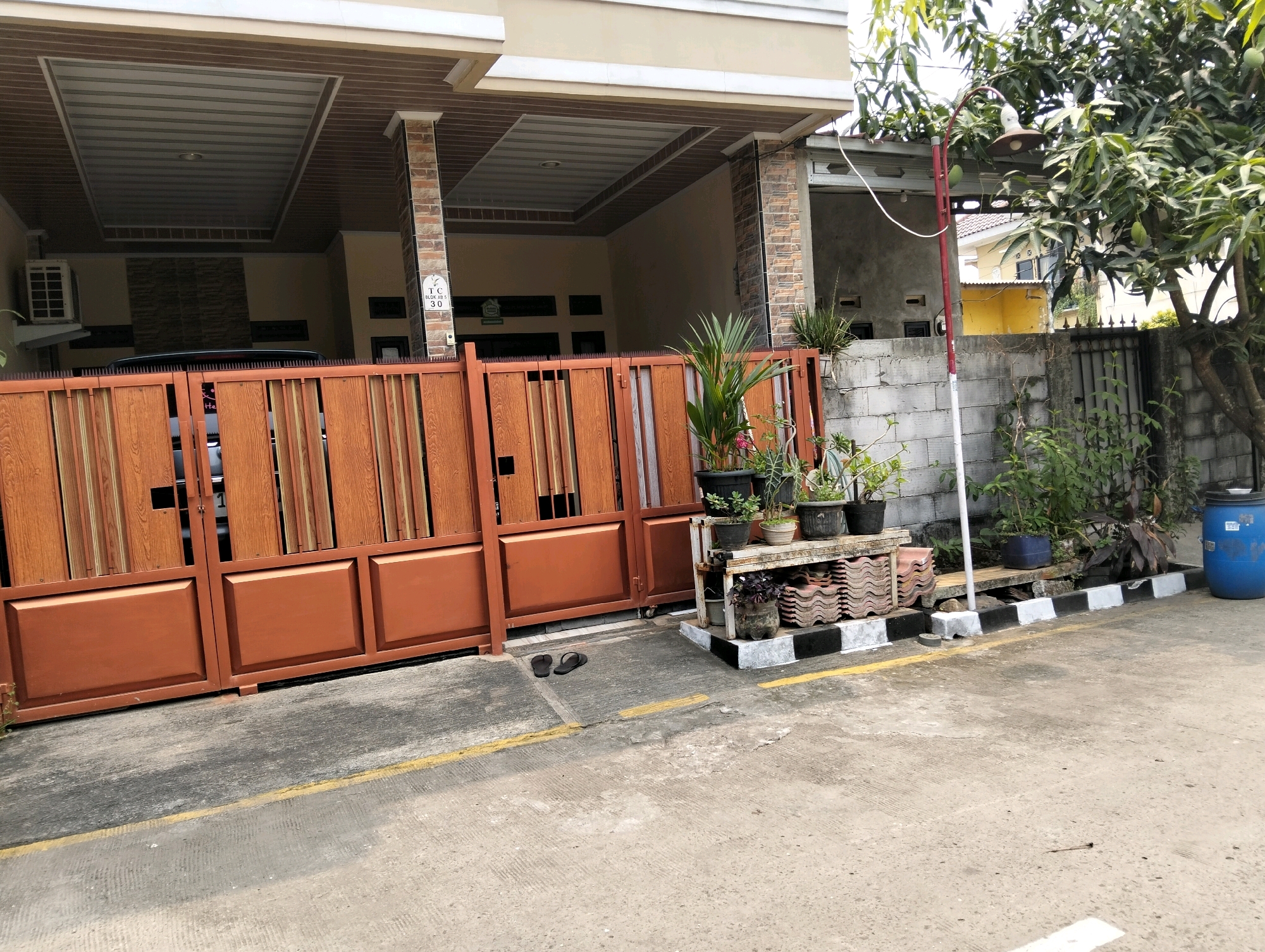 The image depicts the exterior of a residential property located in Cileungsi, Indonesia. The scene is dominated by a large, orange metal gate with vertical wooden panels and decorative rectangular indentations. To the left of the gate, an outdoor air conditioning unit is visible on the exterior wall of the house. The house itself features a beige facade with brown and brick-patterned columns supporting an overhang. Behind the gate, a dark-colored vehicle is partially visible.
In front of the gate and to the right, a collection of potted plants sits on a multi-tiered shelf made of concrete blocks and what appear to be stacked roof tiles. A blue plastic barrel is situated to the far right of the frame, next to a grey cinder block wall. A street lamp with a red pole and a light fixture is positioned to the right of the wall, partially obscured by a large tree with green leaves and visible fruit. The ground in front of the property is paved with concrete and marked with yellow and white lines. A pair of black flip-flops rests on the concrete near the gate. The environment suggests a sunny day in a residential area.