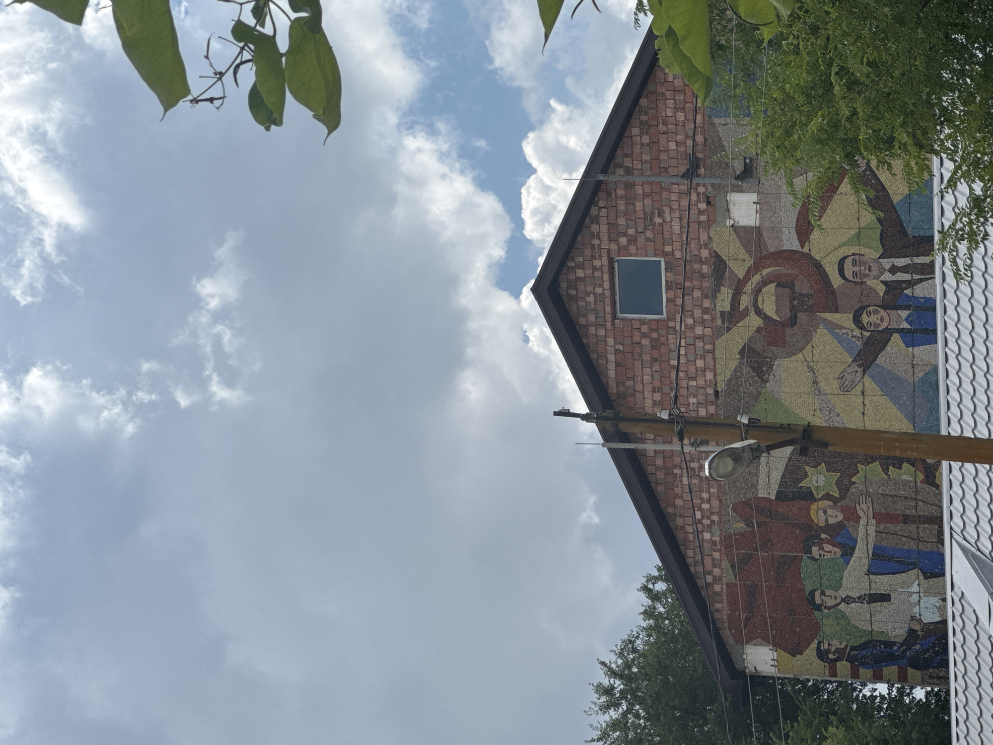 The image shows a mosaic mural on the side of a brick building in Mayak, Russia. The mural depicts a stylized representation of Soviet era propaganda, featuring figures in formal attire, a red flag, and symbols such as a hammer and sickle. The building is topped with a dark-colored gable roof and has a small rectangular window on its upper level. Trees are visible on either side of the building, and a telephone pole with a light fixture stands in front of the mural. The sky above is partly cloudy.