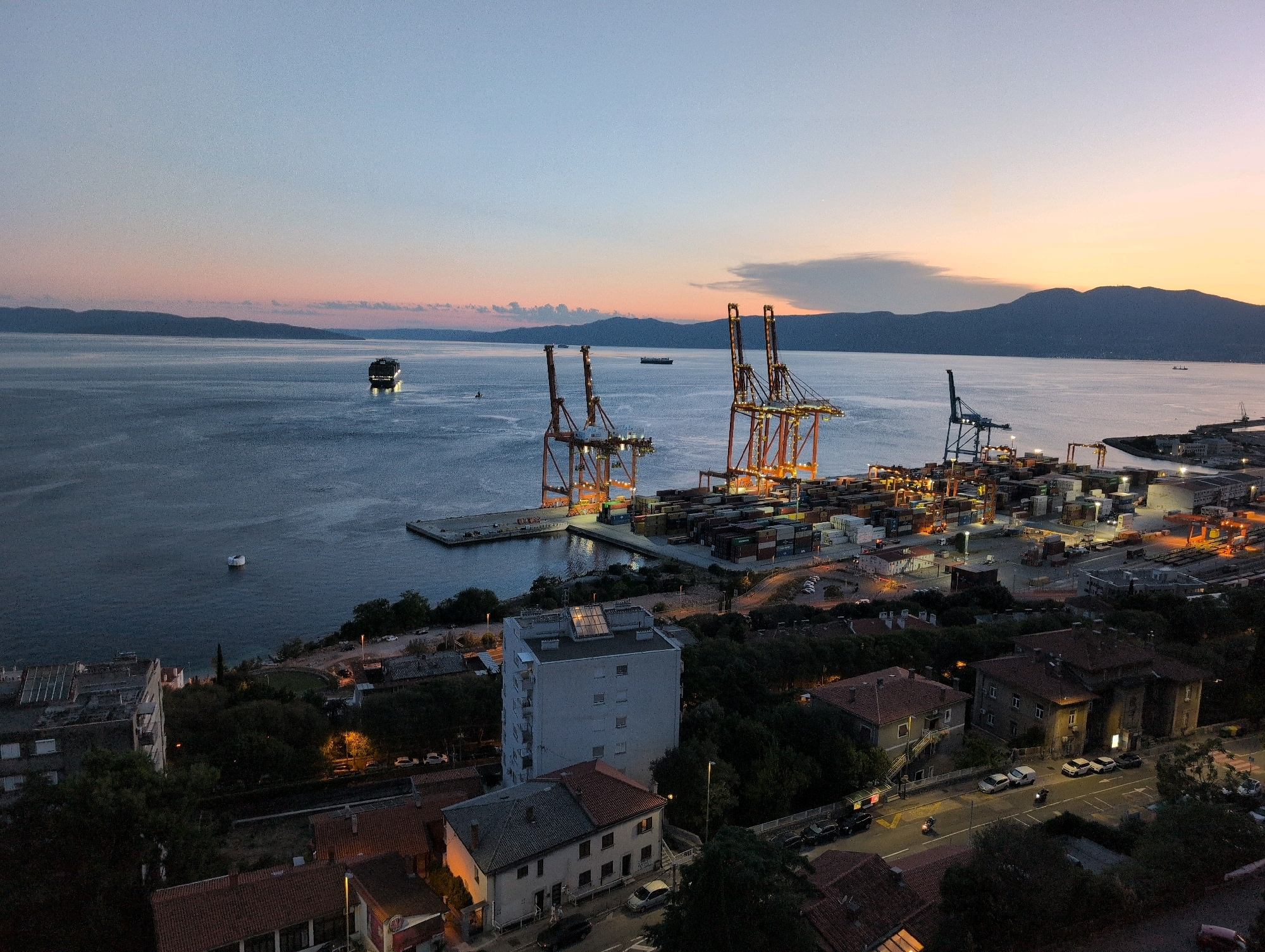 This aerial, wide-angle photograph captures a twilight scene in the city of Rijeka, Croatia, overlooking the Adriatic Sea. The sky is painted with soft hues of blue and orange, transitioning from dusk to night. Dominating the mid-ground is a bustling port complex, illuminated by artificial lights. Several large, orange shipping cranes stand tall, silhouetted against the horizon, with stacks of shipping containers neatly arranged below them. The sea, with its calm surface reflecting the dim light, stretches towards distant, hazy mountains. A large ship is visible in the distance, its lights twinkling on the water. In the foreground, residential buildings with red-tiled roofs are nestled among trees, with streetlights illuminating a road where several cars and two motorcycles are parked or in motion. One white building stands out among the others. The overall atmosphere is one of quiet activity at a coastal industrial and residential area during the evening.