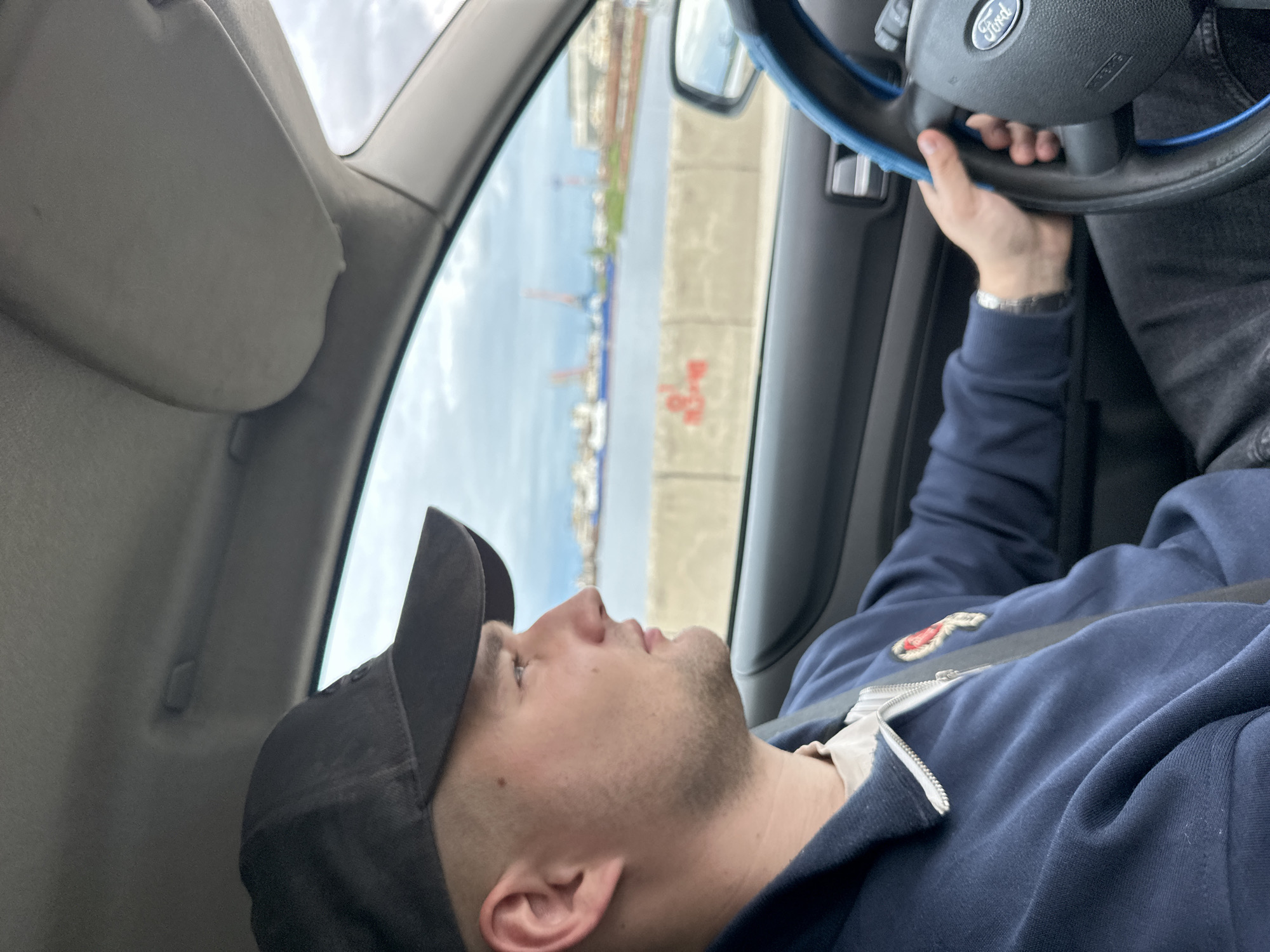 A medium, canted, indoor shot shows a person in a car with their hand on the steering wheel.
The person is a light-skinned man wearing a dark-colored baseball cap, a blue zip-up hooded jacket, and a seat belt. A metal watch is visible on his left wrist. His head is tilted back, and he is looking up and out of the car window. His mouth is slightly open.
The car is a Ford. The Ford logo is visible on the steering wheel. The interior is grey. The car is moving; the visor is down, and a body of water and industrial structures on the shore are seen through the car window. Red graffiti is on a concrete wall that separates the car from the shore. The photograph was taken in the city of Chaikovsky, Russia.