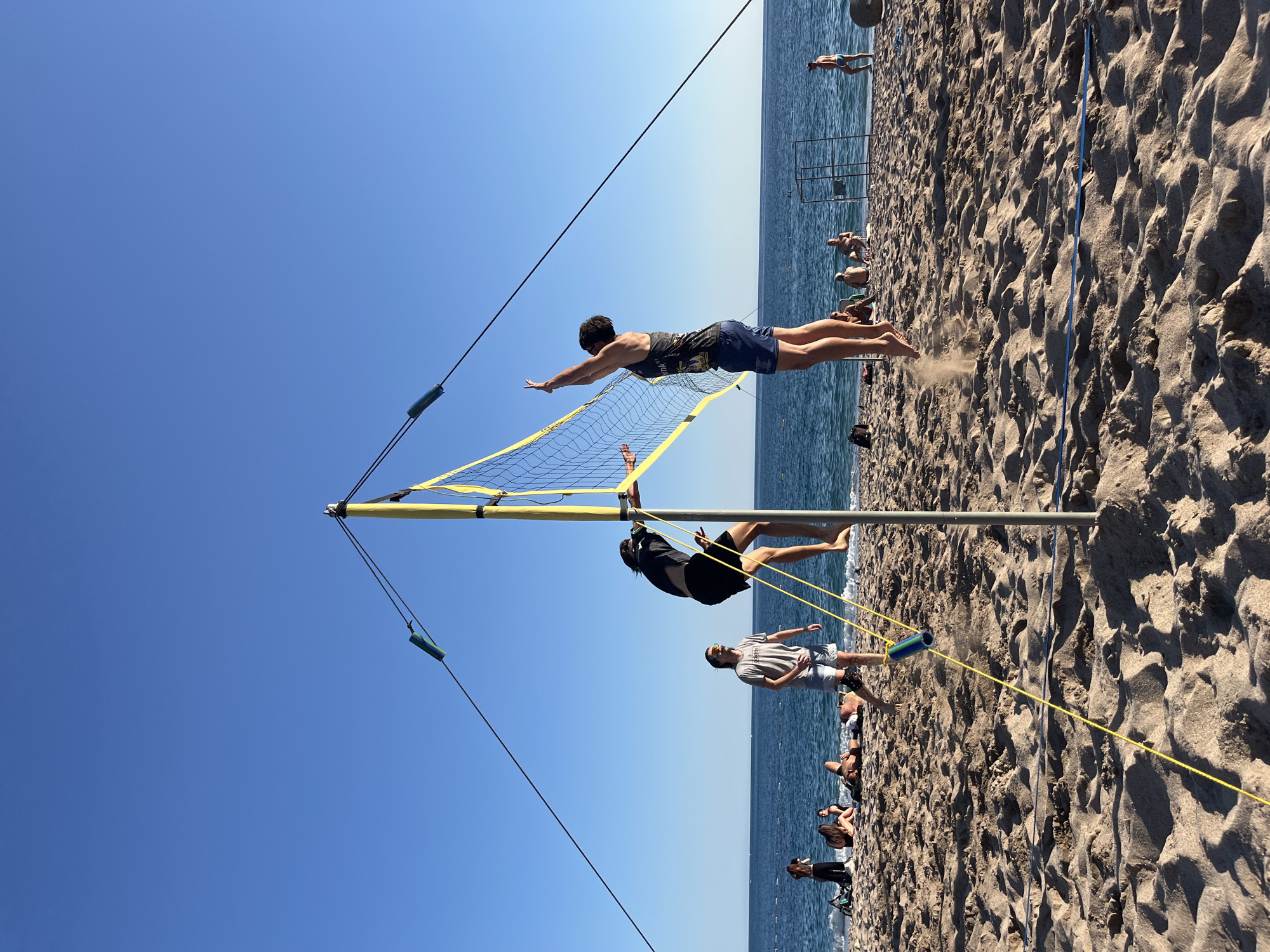 Two young men are playing beach volleyball on a sandy beach in Zankovići, Montenegro, with the Adriatic Sea and a clear blue sky in the background. The player on the right is in mid-air, jumping to spike the ball. He is wearing a black tank top and blue shorts. The player on the left is climbing the volleyball net pole, reaching for the ball. He is wearing a black t-shirt and black shorts. Several other people are visible in the background, relaxing on the beach and swimming in the sea. A volleyball net is set up, and yellow ropes are tethered to the poles, stabilizing the net. The sand is light brown and appears dry, with some footprints visible. The water is a deep blue, and gentle waves are breaking on the shore. The sun is shining brightly, casting shadows on the sand.