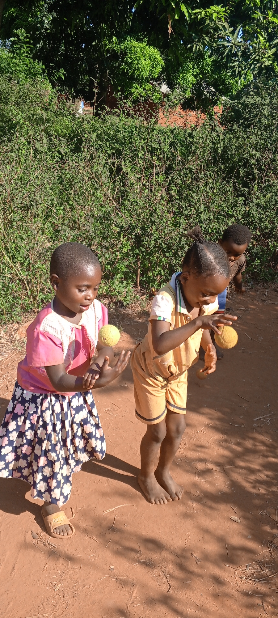 In an outdoor scene, likely Ngalula, Democratic Republic of the Congo, based on context clues such as clothing and environment, three children are present on reddish-brown soil, possibly a yard or unpaved road. In the foreground, two girls are actively engaged in playing with yellow spherical objects, possibly balls or toys. The girl on the left is wearing a pink top with a floral print skirt and has a sandal on one foot. The girl on the right is wearing a yellow shorts outfit with striped trim and is barefoot. A third child is visible in the background, partially obscured. Dense green foliage and trees form the backdrop. The lighting suggests it is daytime with strong sunlight casting shadows.