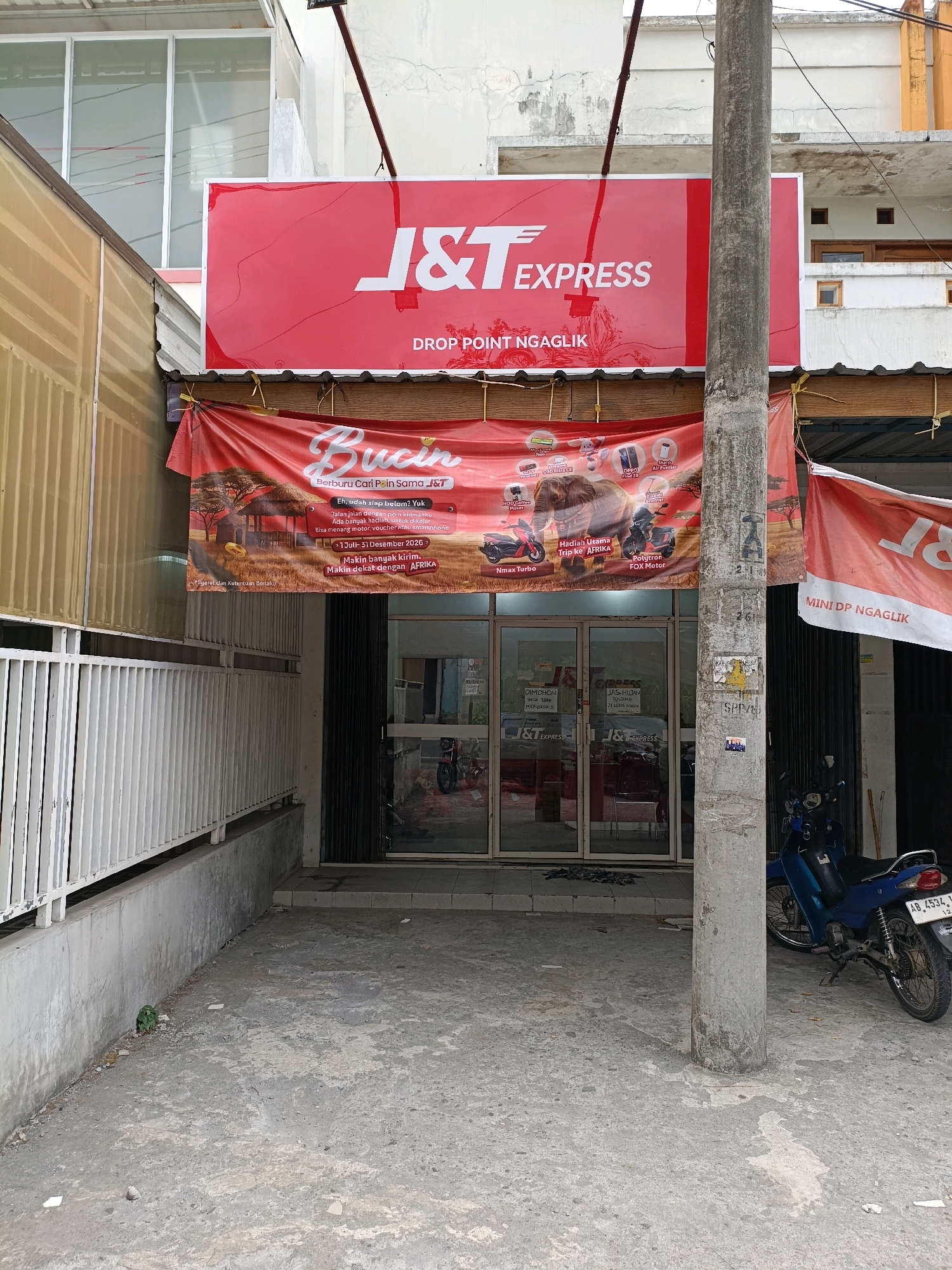 The image shows the exterior of a J&T Express drop point located in Ngaglik, Indonesia. A large red sign with the J&T Express logo and "DROP POINT NGAGLIK" is prominently displayed above the entrance. A red banner with promotional text and images of prizes is hung below the sign. The entrance consists of glass double doors, with the J&T Express logo visible on them. To the right of the entrance, a blue motorcycle is parked. The building appears to be a storefront with a concrete facade and a corrugated metal roof. The ground in front of the building is paved with concrete. A utility pole stands in front of the entrance, obscuring part of the view. The overall environment suggests an urban or semi-urban commercial area.