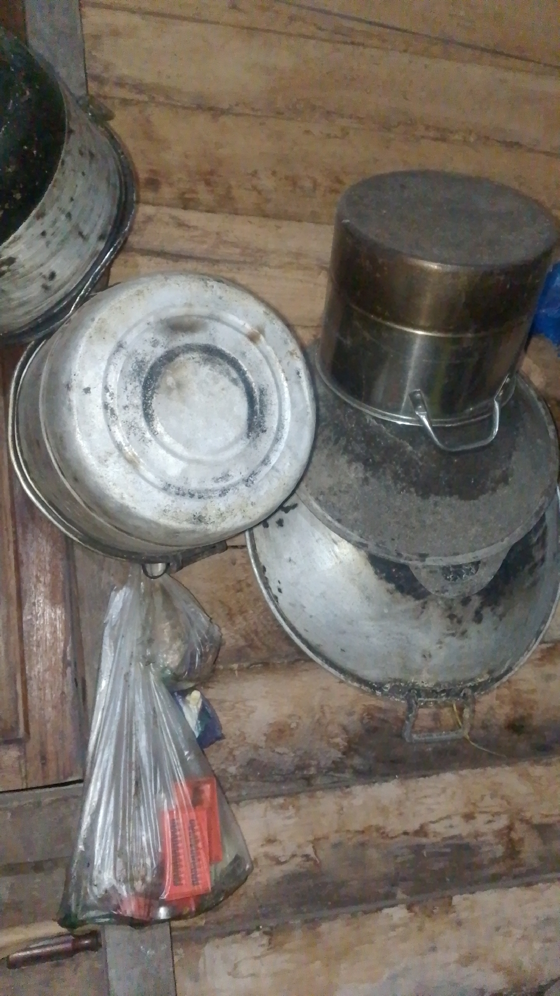 The image depicts a collection of worn, metallic kitchenware and a plastic bag filled with items, possibly food supplies, hanging from a hook. The cookware includes what appears to be pots and pans, some stacked, and one larger, cylindrical container. The objects are placed against a backdrop of rough, wooden planks, suggesting an unrefined or rustic setting. A portion of a knife handle is visible in the lower left corner. The overall scene conveys a sense of practicality and potentially scarcity, with objects showing signs of use and age. The lighting is dim, and the image quality is moderate, with some graininess. There are no people visible in the image.