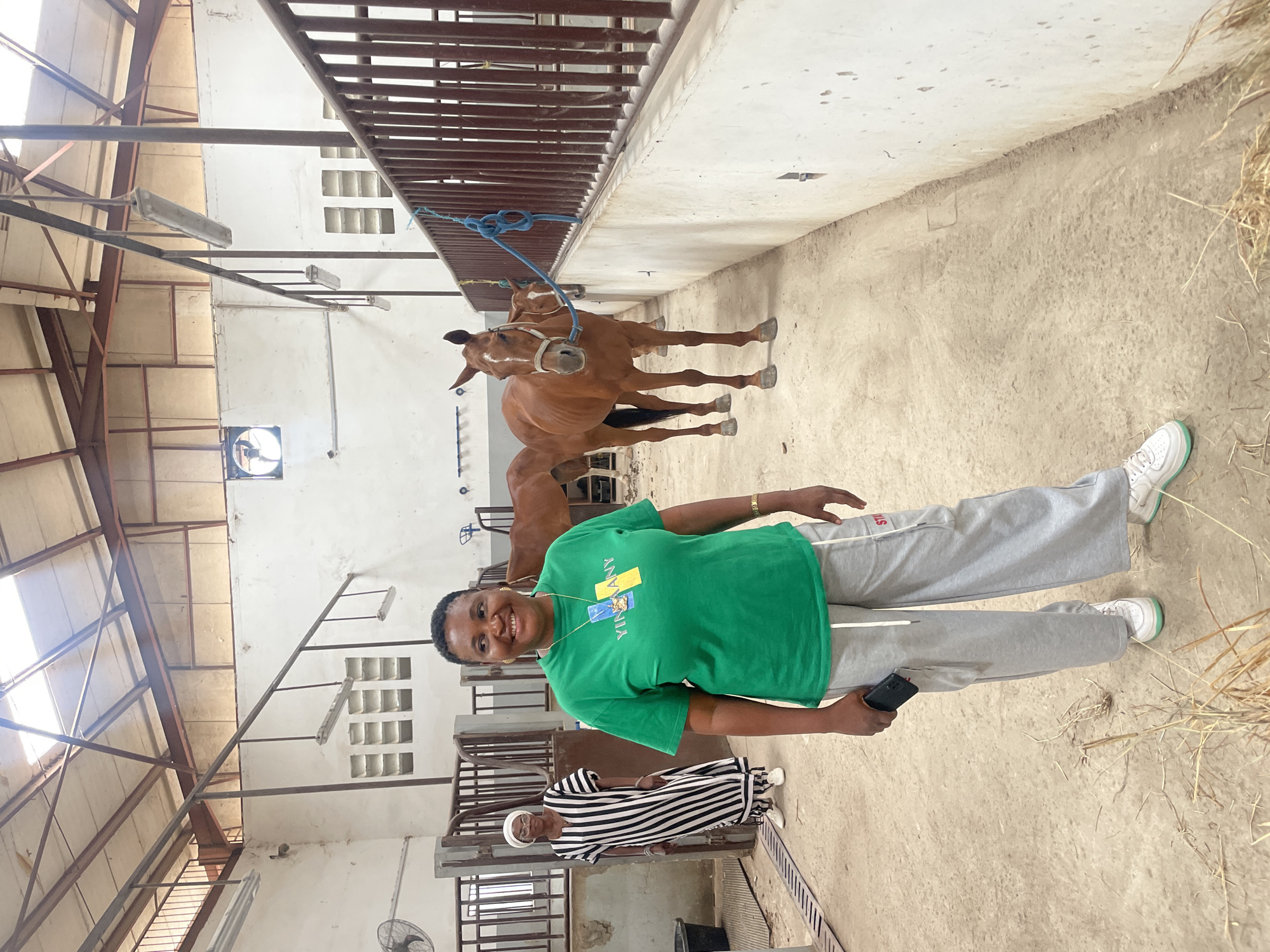 The image shows a woman in a green t-shirt and grey sweatpants standing inside a stable in Hayin Banki, Nigeria. She is smiling and holding a phone in her left hand. Behind her are two brown horses in their stalls, secured with blue ropes. In the background, another person wearing a striped dress and white head covering stands in the adjacent stall. The stable has white walls, metal stall dividers, and a concrete floor. There is hay scattered on the floor near the woman. The stable structure shows exposed metal beams and a fan mounted near the ceiling.
