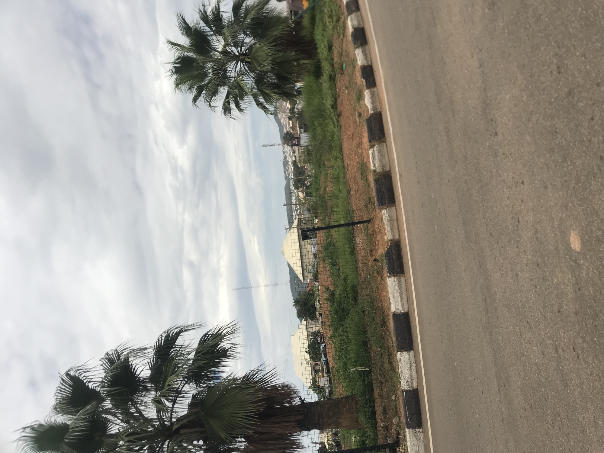 The image depicts a roadside scene in Rafin Par, Nigeria, under a predominantly overcast sky. The foreground features a paved asphalt road, bordered by a curb painted with alternating black and white segments. A white line runs along the edge of the road, separating it from the curb.
Behind the curb, a strip of reddish-brown soil supports sparse green grass and weeds. A black wire mesh fence, held by black metal posts, extends across the midground. Two large palm trees are prominent; one on the far left, partially cropped, with a thick, dark trunk and drooping green fronds, and another smaller palm tree positioned towards the right side of the frame.
Beyond the fence, two buildings with light-colored, possibly cream or white, pyramidal roofs are visible. The building on the left appears to have an antenna or satellite dish on its roof. Further into the midground, green vegetation and patches of reddish-brown earth are present.
The background reveals a distant town or city, characterized by numerous low-rise structures clustered against a backdrop of rolling hills or mountains. A tall, slender communication tower stands visible on the horizon. A partial figure of a person, wearing a yellow garment, is barely visible on the far right edge of the frame.