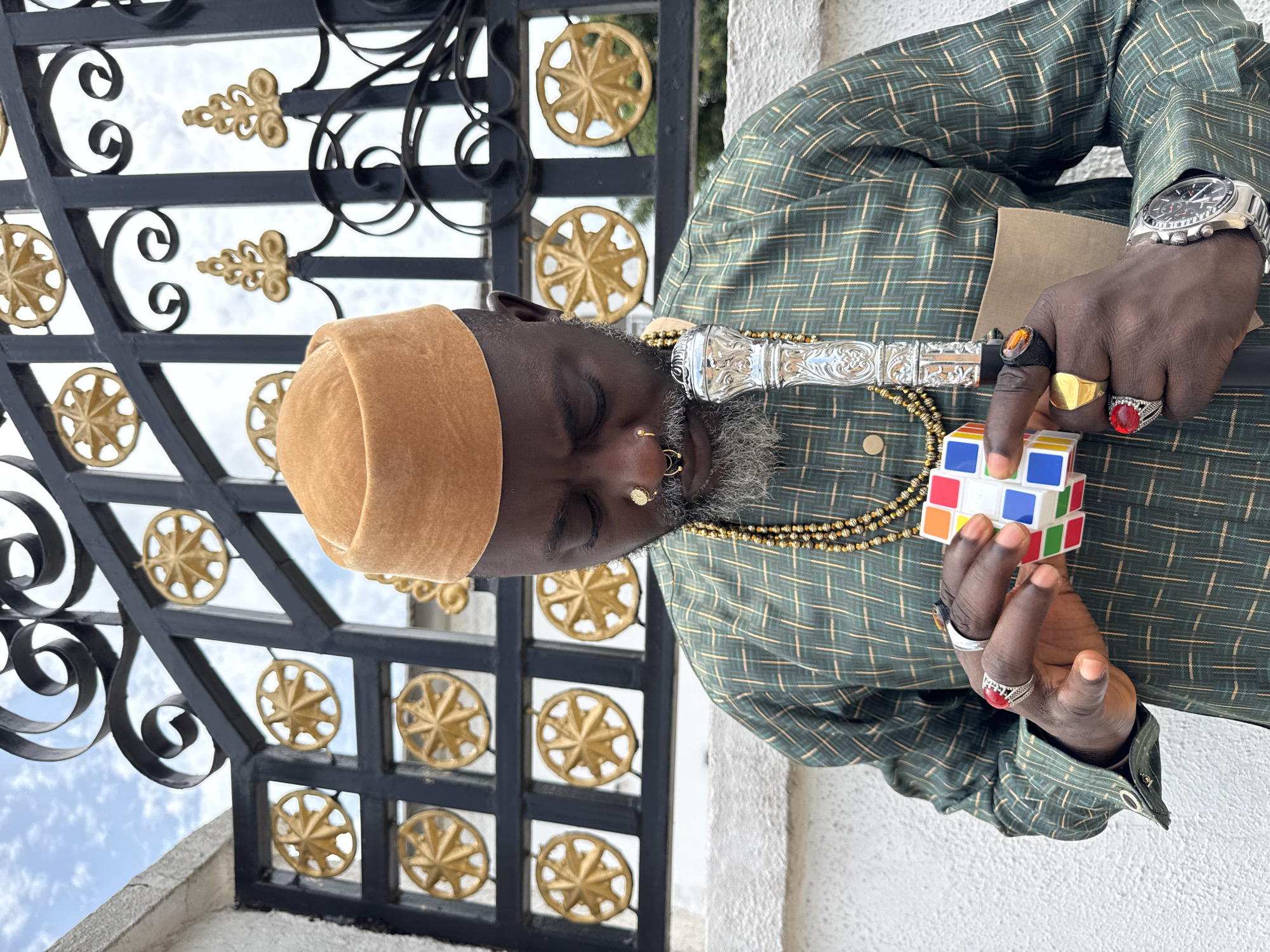 An adult male stands outdoors in Ado, Nigeria, positioned before a black wrought-iron gate with golden star-like and scrollwork decorative elements, against a white textured wall and a cloudy sky. He is observed wearing a light brown velvet cap, a green plaid tunic, and multiple gold-colored bead necklaces. His face features a grey beard and two golden nose rings. With his gaze directed downwards, he actively manipulates a multi-colored Rubik's Cube using both hands. His hands are adorned with numerous rings, including a large red-stoned ring on his left index finger and a black-banded amber-stoned ring on his right middle finger. A silver-colored watch with a black face is visible on his left wrist. He holds an ornate silver-topped staff or cane in his right hand, beneath which a light brown paper or book is partially visible.