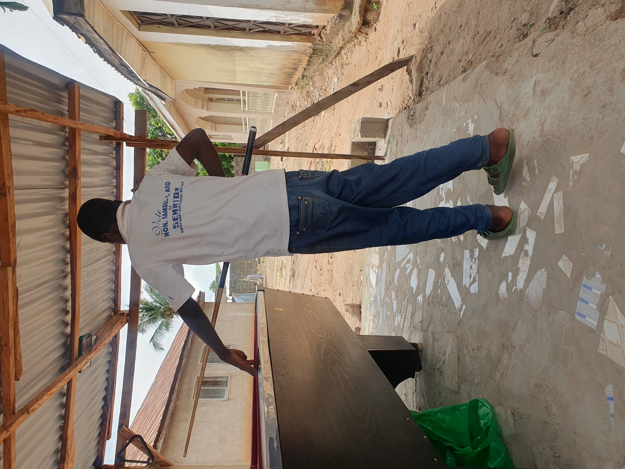 A young Black male wearing a white t-shirt and blue jeans stands near a wooden and metal billiard table. He is outdoors, under a corrugated metal roof supported by wooden beams. The billiard table appears to be in a state of disrepair or under construction. The ground is a mix of concrete and dirt, with scattered broken tiles. In the background, there are buildings with cream-colored walls and tiled roofs, typical of residential areas in Lokoja, Nigeria. Palm trees and other vegetation are visible, suggesting a tropical or subtropical climate. The overall scene depicts an informal setting, possibly a backyard or a community area, where a game or recreational activity is taking place or being set up.