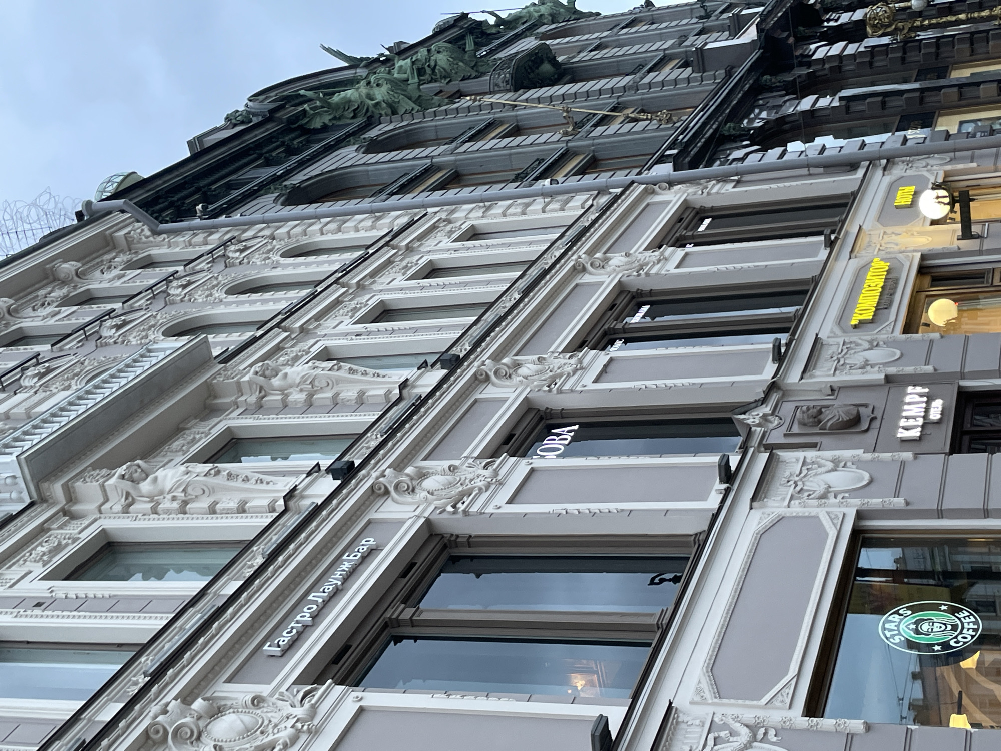The image depicts the architectural details of a building located in Saint Petersburg, Russia. The building features ornate, classical-style facades with decorative moldings, sculptures (including reclining figures), and detailed window frames. There are various storefront signs visible, including "Гастро Лаунж Бар" (Gastro Lounge Bar), "BOBA", "KEMPF", and a Starbucks logo. A sign written in Cyrillic script reads "Композитор" (Kompozitor). The building's upper level is adorned with elaborate metalwork and additional sculptural elements. The sky appears overcast.