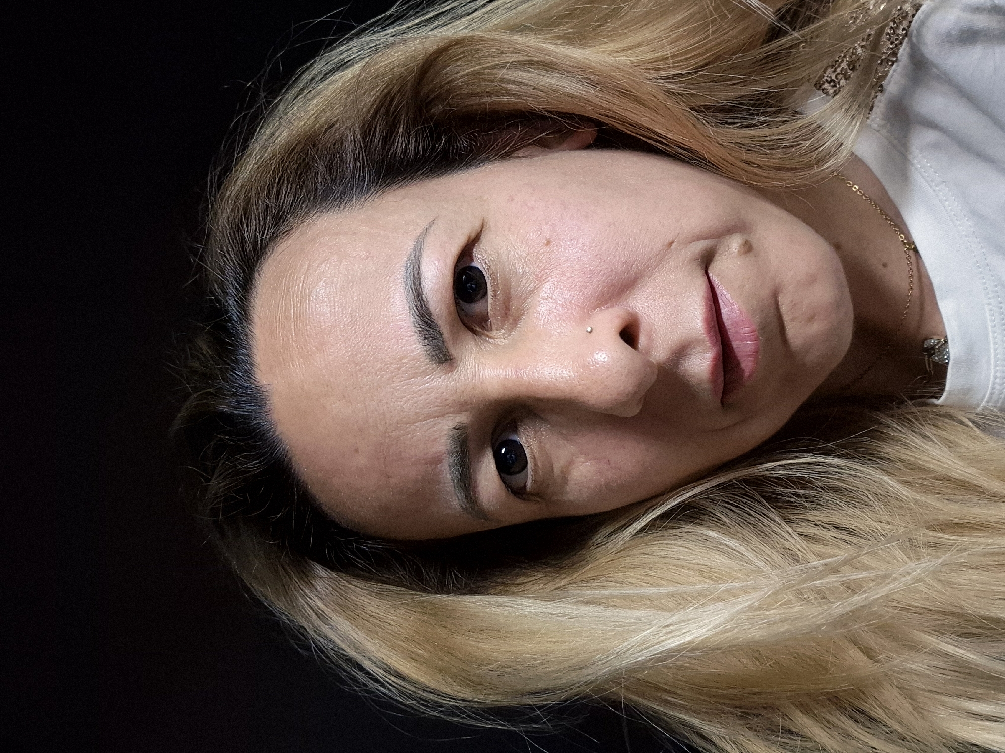 A close-up, head-and-shoulders selfie of a woman with blonde hair and a dark background. She is looking directly at the camera with a neutral expression. Her skin is fair and shows some signs of aging. She has a small mole on her left cheek and a nose stud. Her hair is long and wavy, framing her face. She is wearing a white t-shirt and a gold necklace. The lighting is somewhat harsh, casting shadows on her face. The image appears to be taken indoors, with no discernible urban environment.