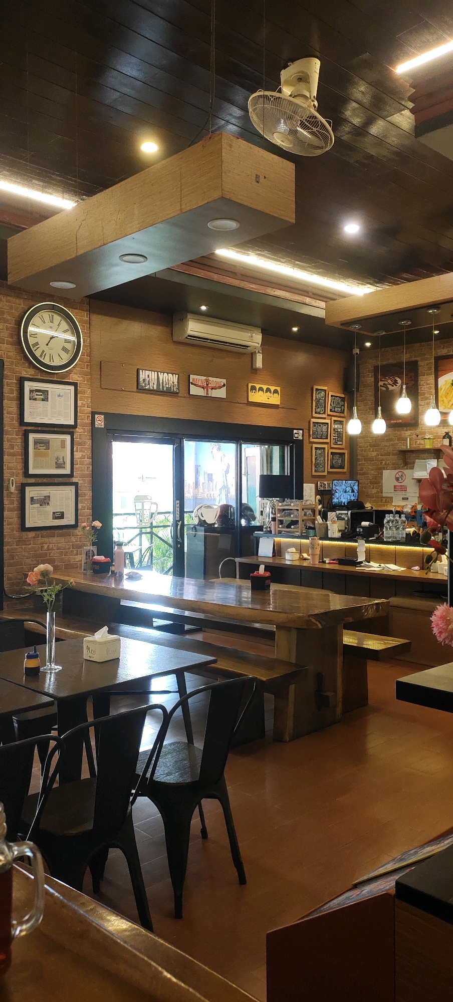 The image depicts the interior of a cafe in Nganguk, Indonesia. The cafe features a rustic and modern design, with wooden furniture, exposed brick walls, and a dark wooden ceiling. Long, communal wooden tables with matching benches are positioned throughout the space. Black metal chairs are also present.
On the brick wall, a large round clock with Roman numerals hangs prominently. Adjacent to the clock are framed pieces of what appear to be newspaper clippings. Further along the wall, several framed artworks and signs are displayed, including one that says "NEW YORK" and another featuring a silhouette of a runner labeled "WAYNE ROONEY." A cluster of framed chalkboards with various designs adds to the decor.
The ceiling is adorned with a white ceiling fan and modern light fixtures, including linear fluorescent lights and circular recessed lights. A central, large, wooden beam structure runs horizontally across a portion of the ceiling, integrated with lighting.
In the background, a counter area is visible, with shelves stocked with bottles and various items. A computer monitor sits on the counter, displaying an image. Through large glass doors in the background, an outdoor seating area with greenery can be seen.
Several small objects are placed on the tables, including a vase with flowers and tissue boxes. The lighting in the cafe is a mix of natural light coming from the outdoor area and artificial lighting from the ceiling fixtures, creating a warm and inviting ambiance. No people are actively visible in the scene.