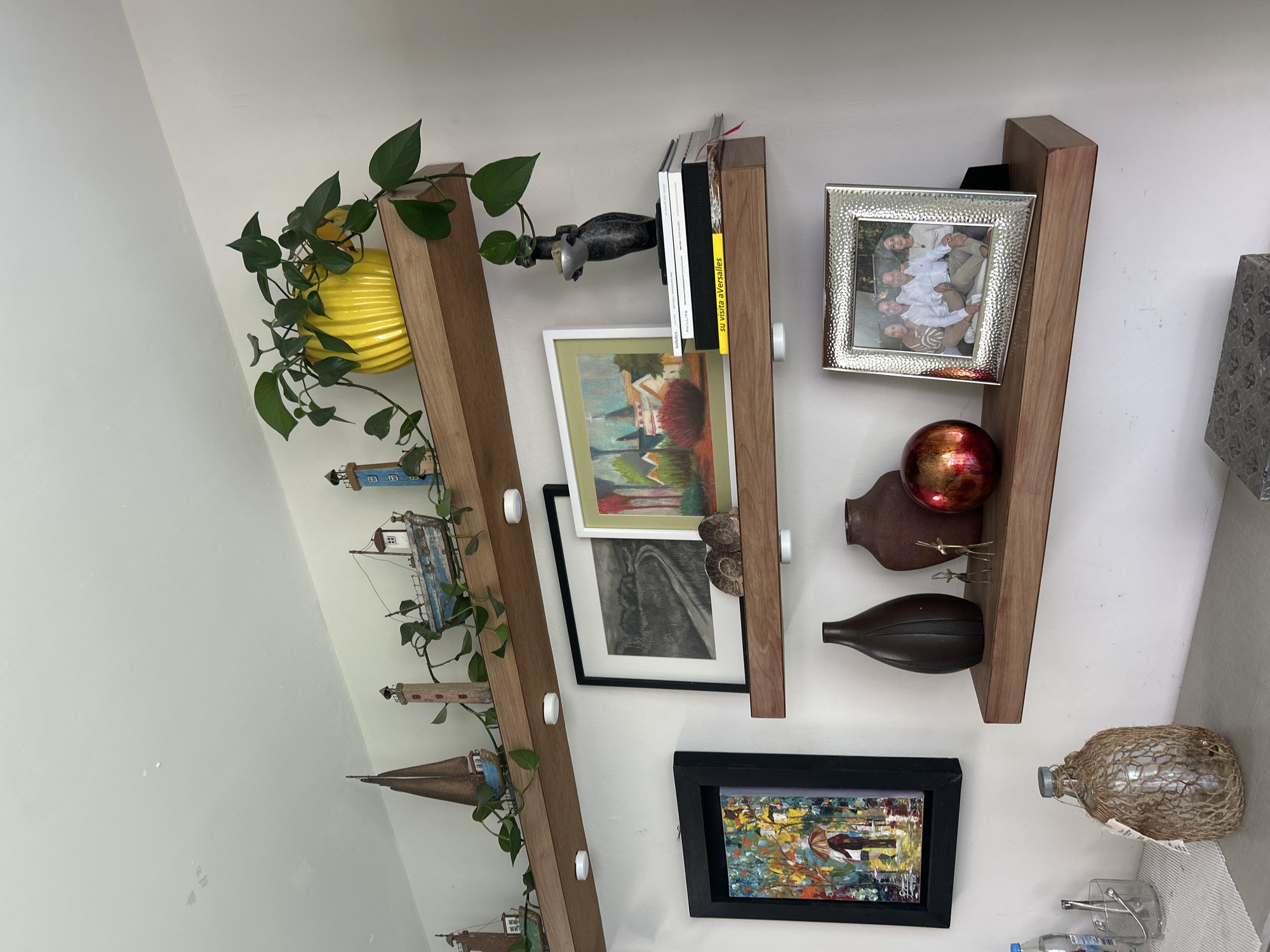 The image shows three vertical wooden shelves mounted on a white wall. Each shelf is decorated with various items. The top shelf holds a yellow ridged pot with a trailing green plant, a black decorative object resembling a human hand, and several books, including one with a yellow spine.
The middle shelf displays a framed painting with a white matte, a black and white artwork in a black frame, two small brown decorative vases, a metallic red sphere, and a small gold ornament.
The bottom shelf features a framed painting with a black frame, and a glass bottle wrapped in twine.
The location of the image is Barrio de Nuevo León, Mexico.