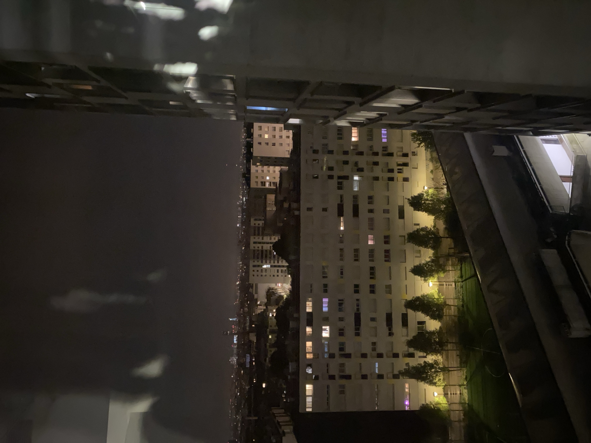The image captures a nighttime view in Paris, France, specifically within a densely populated urban area characterized by high-rise apartment buildings. The perspective is from an elevated position, likely within or between buildings, offering a glimpse into the urban landscape.
The immediate foreground is dominated by the concrete structure of the building where the photograph was taken, framing the scene. Beyond this, the cityscape unfolds, showcasing a mix of residential buildings, many with illuminated windows suggesting occupancy. These buildings vary in height and architectural style, contributing to the diverse urban fabric.
Visible between the buildings is a green space with trees and lampposts. In the background, there are lights from other buildings and streets. The overall atmosphere is urban, with the interplay of light and shadow emphasizing the depth and complexity of the city at night.