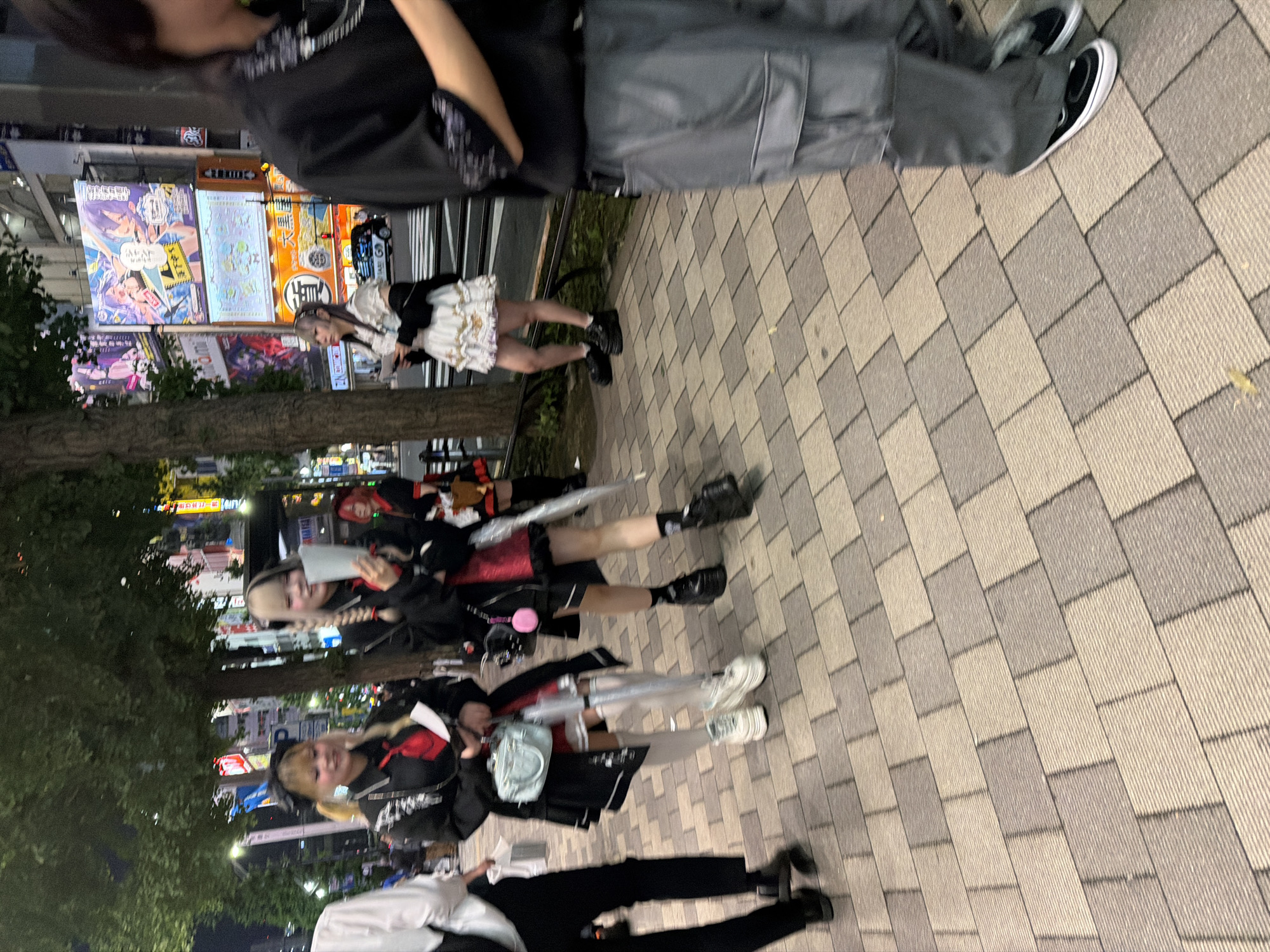 The image depicts a street scene at night in Chiyoda, Japan, likely in an entertainment district, as evidenced by the large illuminated billboards in the background. In the foreground, several individuals are walking on a paved sidewalk. To the left, a person in a white shirt and black pants is walking away from the camera. In the center, two young women in Lolita-inspired fashion are visible. The woman on the left wears a black jacket over a red dress, white thigh-high socks, and platform sneakers. She is holding a light blue handbag and a folded paper. The woman next to her has blonde and black braided hair, a similar black jacket over a red outfit, black socks, and platform boots. She is holding a white paper. Further back, another individual in Lolita fashion is walking away from the camera, and a person in a black shirt and grey cargo pants is walking towards the camera on the right side of the frame. The environment is characteristic of a busy urban street with trees lining the sidewalk and neon lights from surrounding buildings reflecting on the wet pavement.