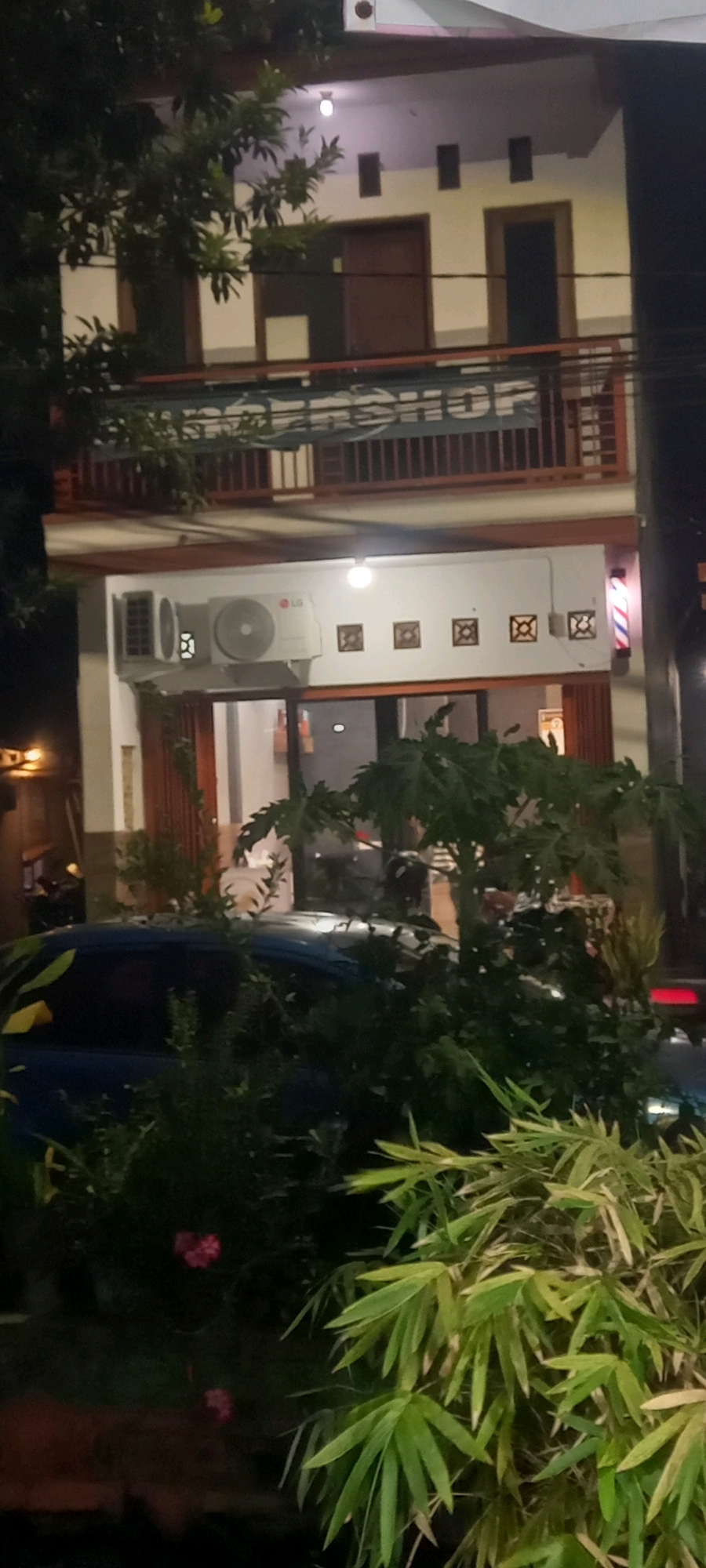 This image depicts the exterior of a building in Sedati Agung, Indonesia, at night. The building appears to be a multi-story structure with a business on the ground floor. On the left side of the ground floor, there is an LG air conditioning unit mounted on the wall. The ground floor also features a glass-paneled door and several decorative square motifs on the wall above it. To the right of this area, a barber pole with red, white, and blue stripes is visible. In front of the building, a blue car is partially obscured by dense foliage. The foreground is dominated by large green leafy plants, with a hint of red flowers and their reflection in the lower left portion of the image. Overhead, there are power lines and a railing. The overall lighting suggests it is nighttime, with artificial lights illuminating the building and its surroundings.