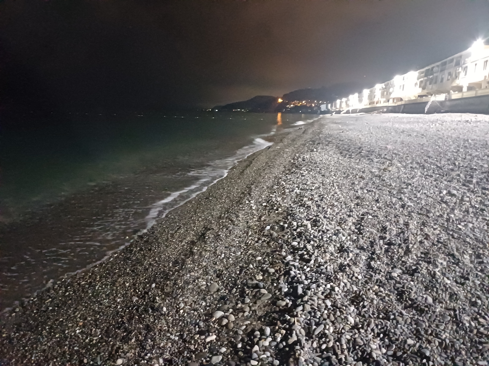 The image captures a nighttime view of a pebble beach along the Black Sea in Sochi, Russia. In the foreground, a broad expanse of grey and dark-toned pebbles extends across the frame, becoming visibly wet and darker closer to the water. A thin line of white foam from gentle waves marks the precise edge where the sea meets the shore.
The midground consists of the dark, calm waters of the Black Sea, reflecting faint light sources. A series of small, bright, horizontally spaced lights are visible across the distant water, consistent with buoys or lights from vessels.
The background on the right features a long, multi-story building or complex running parallel to the coastline. This structure is brightly illuminated by numerous exterior lights, casting a significant glow. To the left of the illuminated building, the coastline recedes into darkness, revealing the faint silhouettes of distant hills or mountains. Scattered yellow and white artificial lights dot the distant landscape, indicating further urban development along the coast. The sky above is dark and appears to be overcast, with a subtle glow near the horizon, likely from light pollution. No individuals or specific actions are visible in the scene.