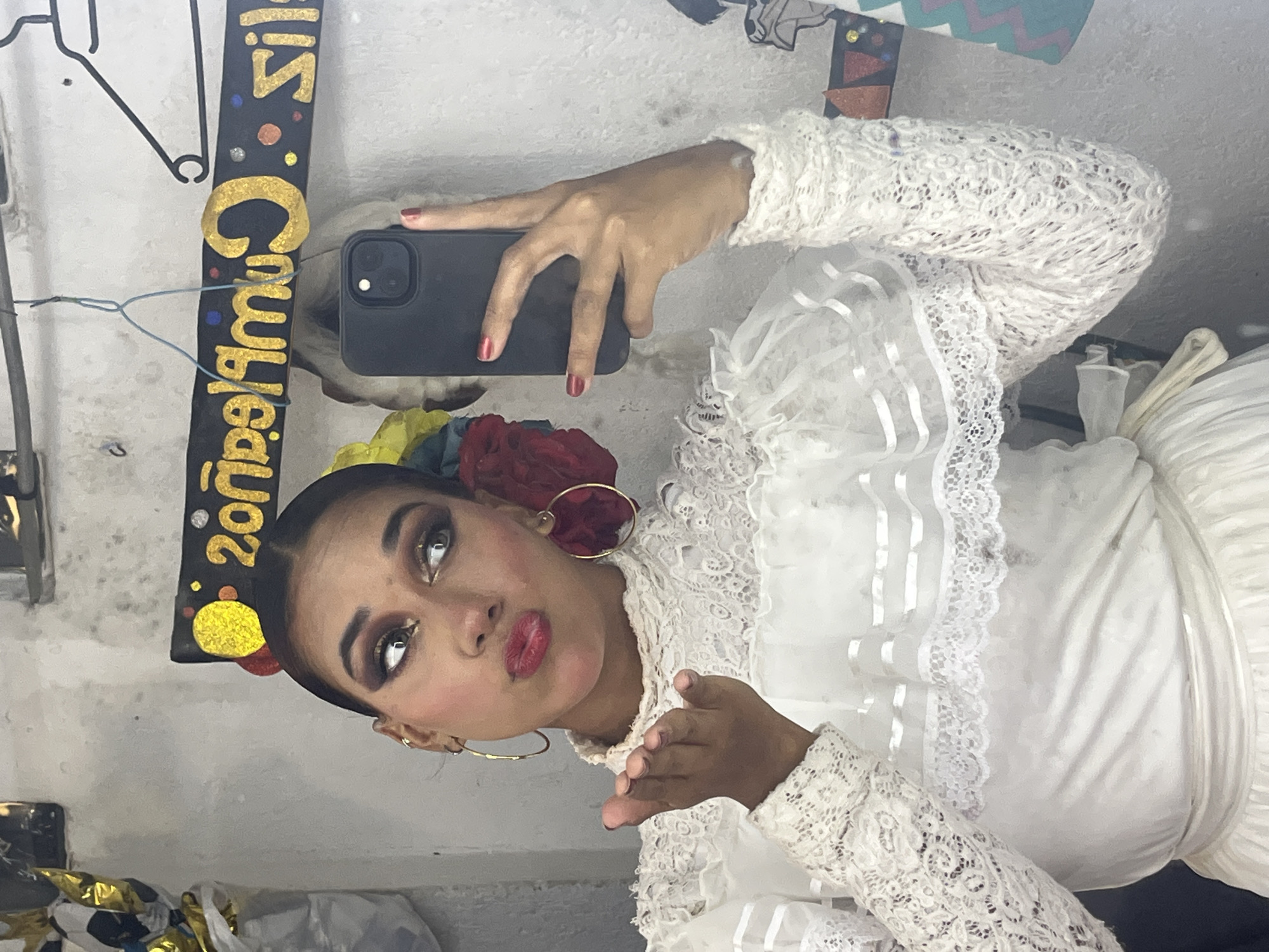 In an indoor setting, a young woman with dark hair pulled back, wearing bold eye makeup and red lipstick, poses for a selfie in a white lace dress. She puckers her lips and holds out her left hand as if blowing a kiss. A black smartphone is held in her right hand, angled to capture her reflection. A black banner with gold lettering that reads "Cumpleaños Feliz" (Happy Birthday) is visible in the background, along with colorful decorations. The wall appears to be textured and light-colored. The overall impression is of a celebratory moment captured in a casual, personal space, possibly a dressing room or a home. The context of Porlamar, Venezuela is not evident from the visual information in the image itself.