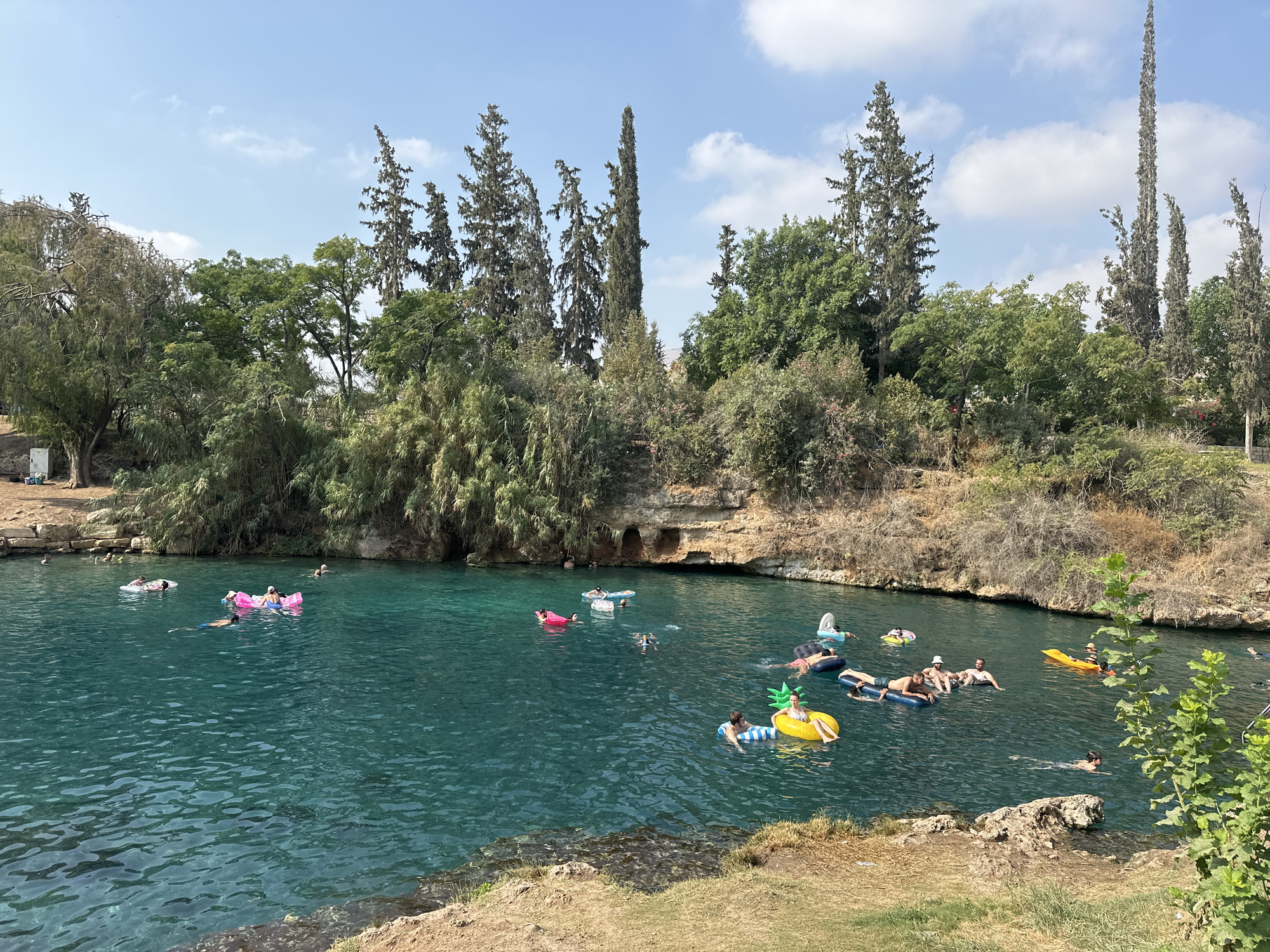 The image depicts a natural spring in Emek HaMaayanot Regional Council, Israel, with a group of people enjoying the water. The spring is characterized by its clear, turquoise water and rocky banks lined with lush green vegetation and tall trees. Several individuals are seen floating on inflatable tubes and mattresses, engaging in recreational activities. Some are swimming or wading in the water, while others are relaxing on their floats. The scene appears peaceful and idyllic, with a clear blue sky and scattered clouds overhead, suggesting a warm and sunny day. The presence of diverse flora and the natural water body point to a well-preserved natural environment within the region.