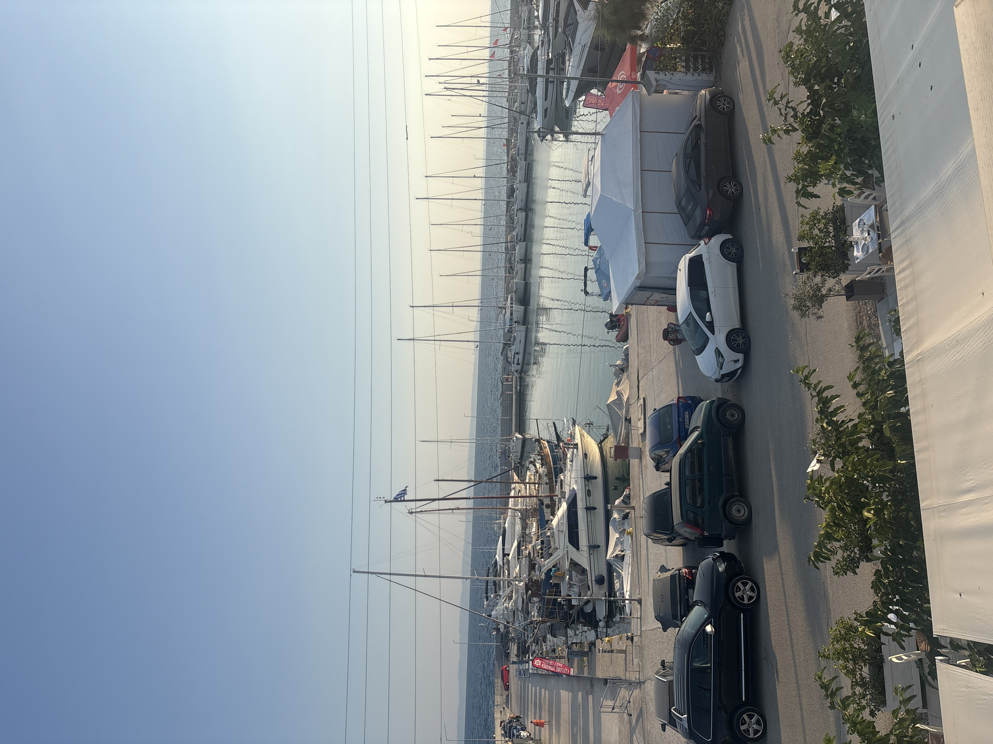 The image depicts a marina in Sithonia Municipal Unit, Greece, under a clear, pale blue sky. The scene is divided into two main areas: the water with docked boats and the land with parked cars and some structures.
In the water, numerous sailboats and motor yachts are moored in rows. The boats vary in size and design, with masts extending high into the air. Some have their sails furled, while others appear ready for sailing. The water is calm, reflecting the boats and the sky.
On the land adjacent to the marina, several cars are parked. These include a black SUV, a dark sedan, a green compact SUV, a white hatchback, and a dark gray sedan. The cars are parked in an asphalt area, some facing the water and others perpendicular to it.
Further inland, there are some temporary structures, possibly tents or canopies, set up on the ground. One white tent is particularly prominent. Some vegetation, including green bushes and small trees, is visible in planters and along the edge of the scene. A few tables and chairs are set up outside, suggesting an outdoor dining area.
A person in an orange shirt is walking along the pier, and another person in darker clothing is visible in the background near the docked boats. The overall atmosphere is one of a peaceful, sunny day at a coastal location.