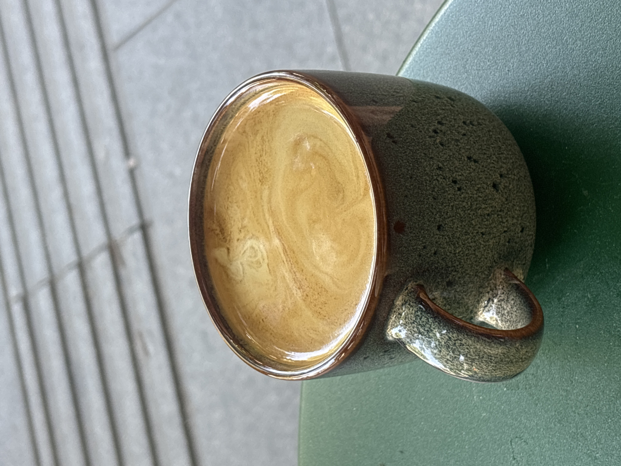 A close-up shot shows a rustic, speckled green ceramic mug filled with creamy, light brown coffee. The coffee's surface exhibits swirling patterns, suggesting it's a latte or cappuccino. The mug has a brown rim and a matching brown handle. It rests on a textured, dark green surface, possibly a table. In the background, out of focus, is a light gray, textured surface with a pattern of parallel lines, resembling outdoor paving stones or a tiled floor. The lighting suggests it is daytime. The overall impression is one of a simple, quiet moment enjoying a coffee, likely outdoors or in a cafe. There is no visible text in the image.