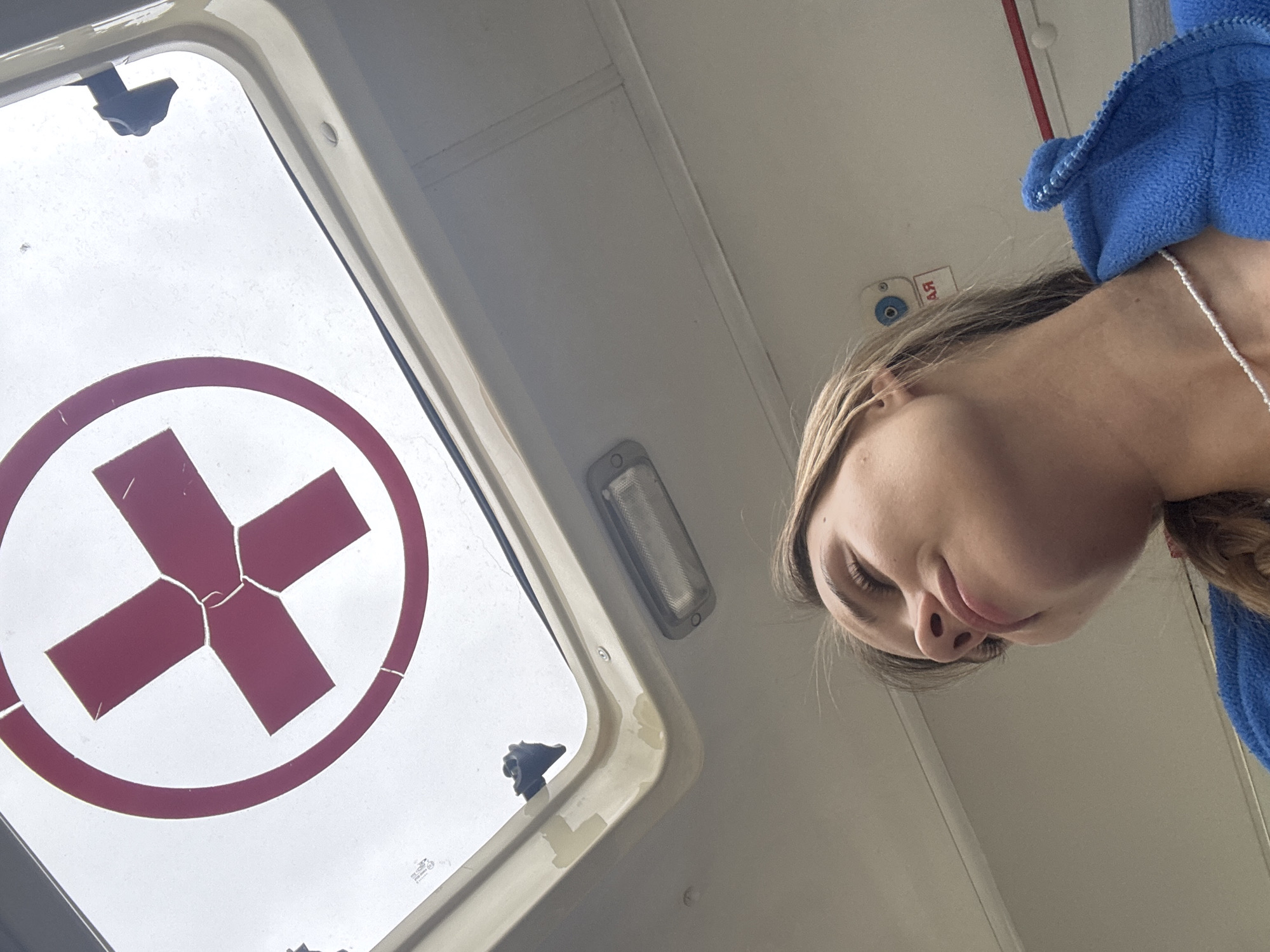 This image depicts the interior of a medical transport vehicle, likely an ambulance, situated in the city of Chaikovsky, Russia. The perspective is a low-angle shot, looking upwards towards the ceiling.
Dominating the upper portion of the frame is an overhead skylight or roof hatch, featuring a large, faded red cross symbol enclosed within a red circular outline. The red material of the cross shows visible cracks and wear. The skylight's frame is a light, off-white or beige color, and a dark, multi-faceted latch mechanism is visible on the upper right side of the frame. Below the skylight, a rectangular, light-colored overhead lamp fixture is mounted on the ceiling.
In the lower half of the frame, the upper body and face of a young female are visible. She is positioned to the left and is looking upwards and slightly to her left, with her eyes appearing partially closed or downcast. Her expression is neutral. She has light-colored hair, possibly blonde or light brown, which appears to be styled into a braid visible on her right side (viewer's right). She is wearing a blue fleece jacket with a visible zipper closure. A thin white beaded necklace is around her neck. The ceiling behind her is a uniform light color, and further to the right, a red horizontal strip with partially visible Cyrillic text ("АЯ") and a small, circular blue object are mounted.
